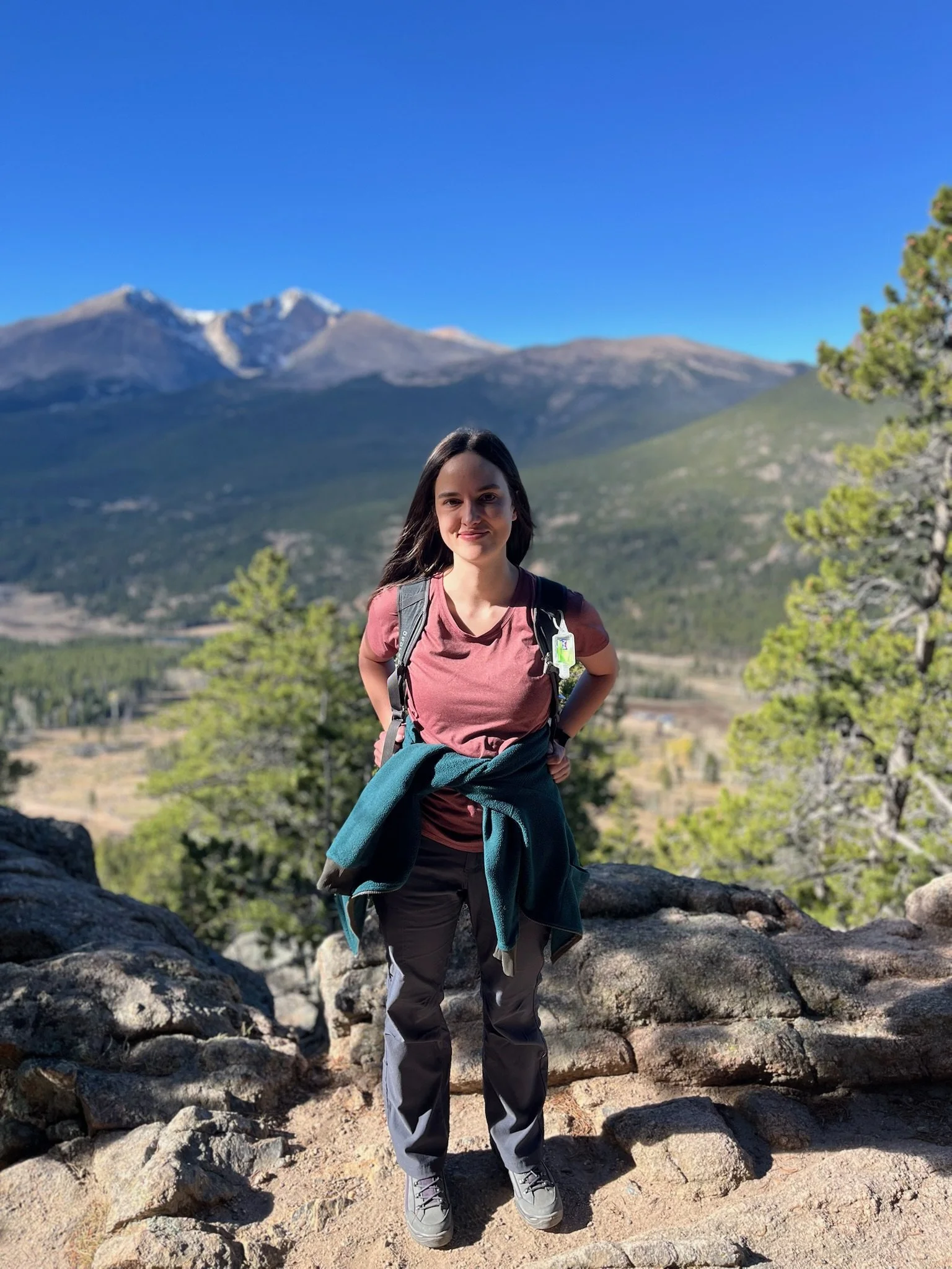 A woman with long dark hair, dressed in hiking clothes, stands on rocks with a mountainous landscape and clear blue sky in the background.
