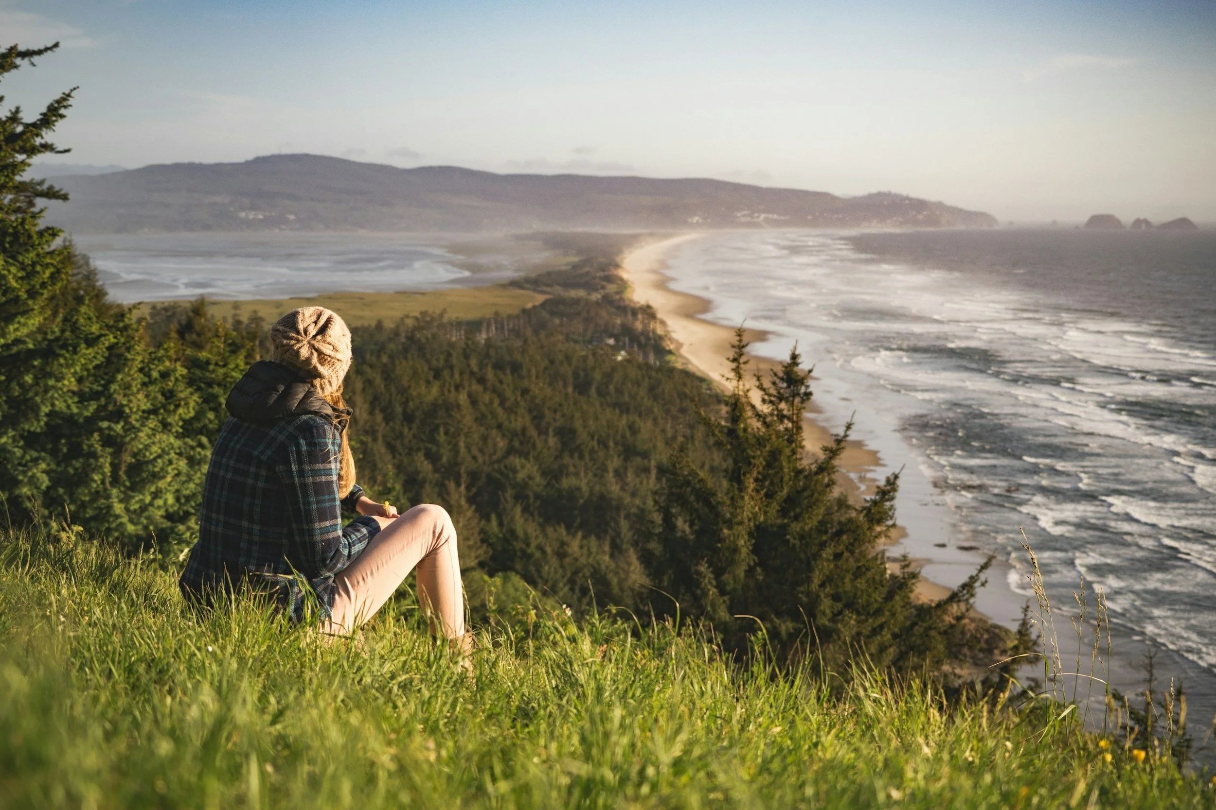 Woman in a beanie and flannel sitting in the grass staring at the ocean with mountains in the distance