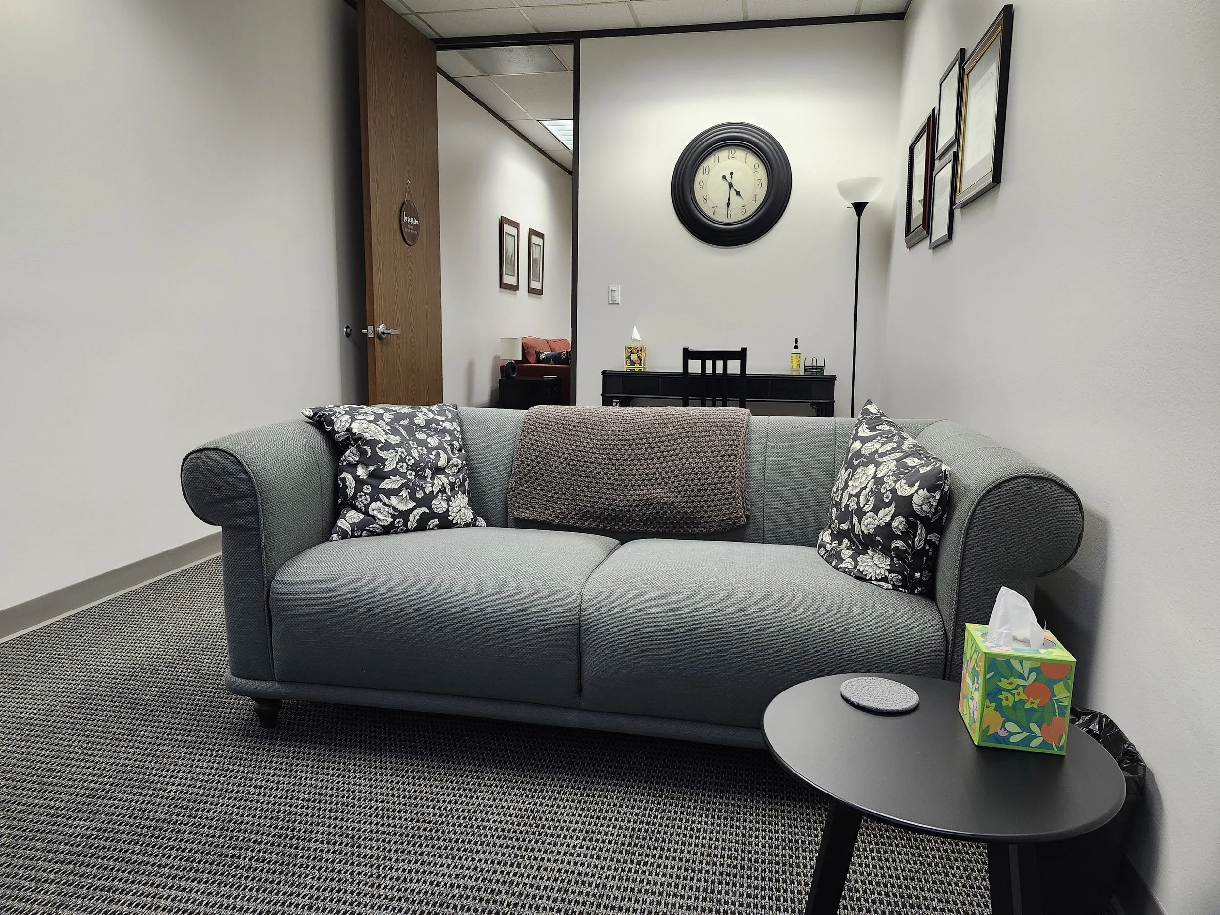 A waiting room with a light blue sofa with two floral-patterned pillows and a brown blanket, a small black side table with a tissue box and coaster, a black desk with a chair, a large wall clock, framed pictures on the wall, a tall floor lamp, and a