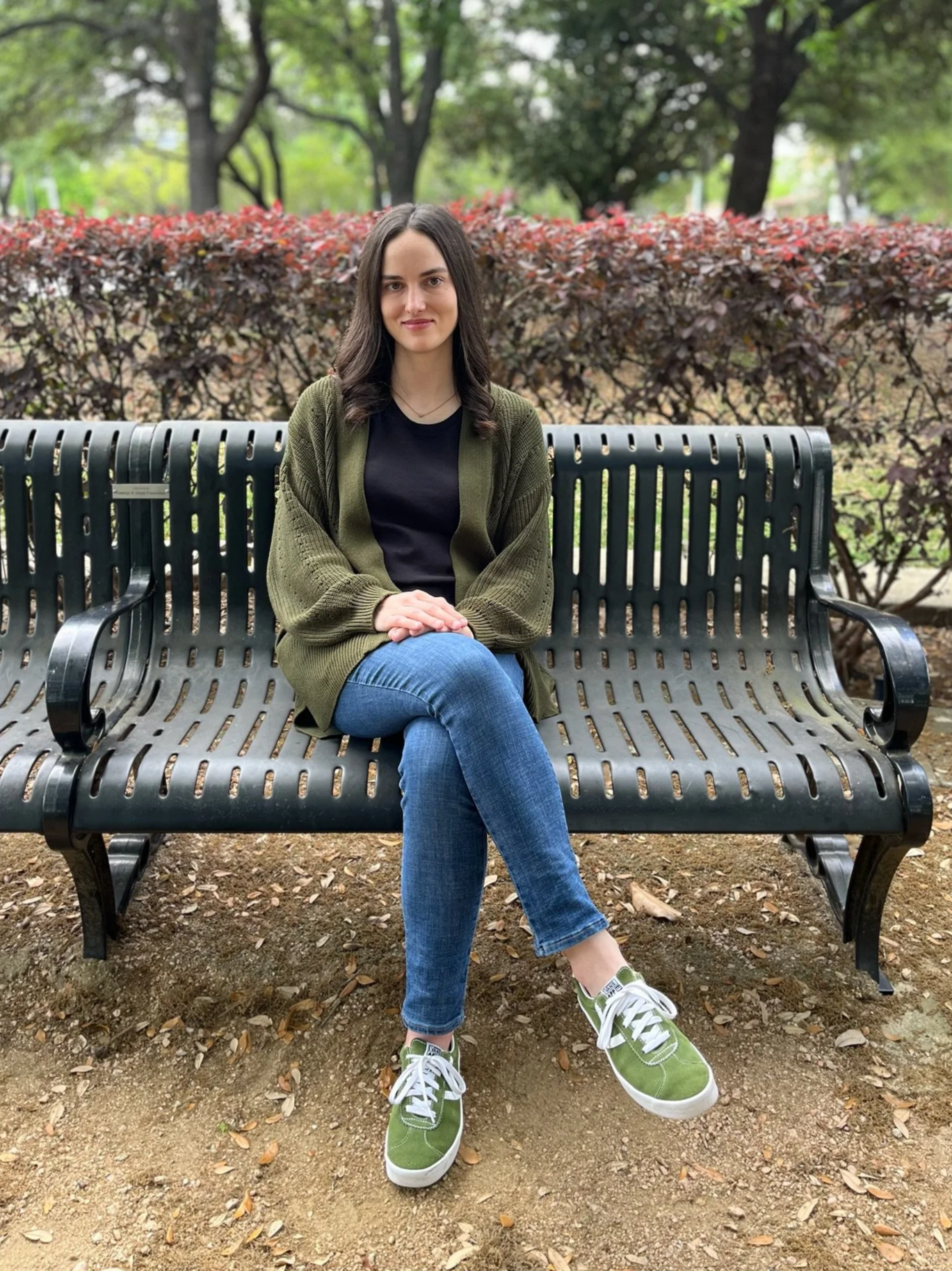 Young woman with long dark hair sitting on a park bench surrounded by greenery and bushes.