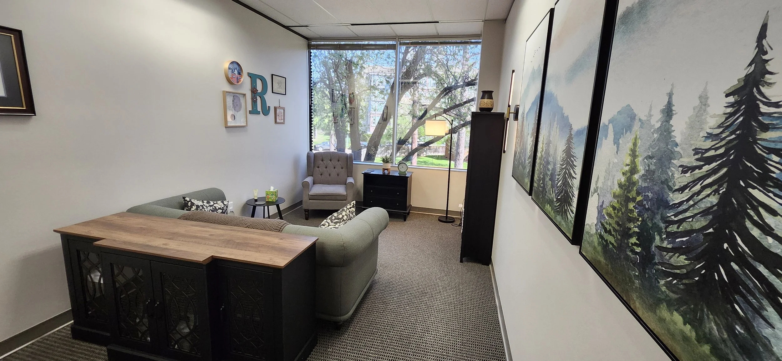 office with a tan armchair near a large window, a green sofa with patterned pillows, black furniture, and forest-themed artwork on the walls.