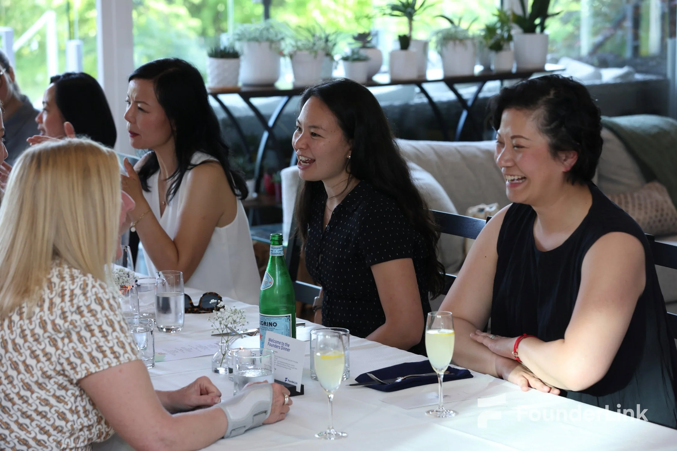 Women sitting at a table with drinks and conversation, smiling and engaged in discussion at a casual gathering or meeting. The scene appears relaxed and friendly, with natural light and plants in the background.