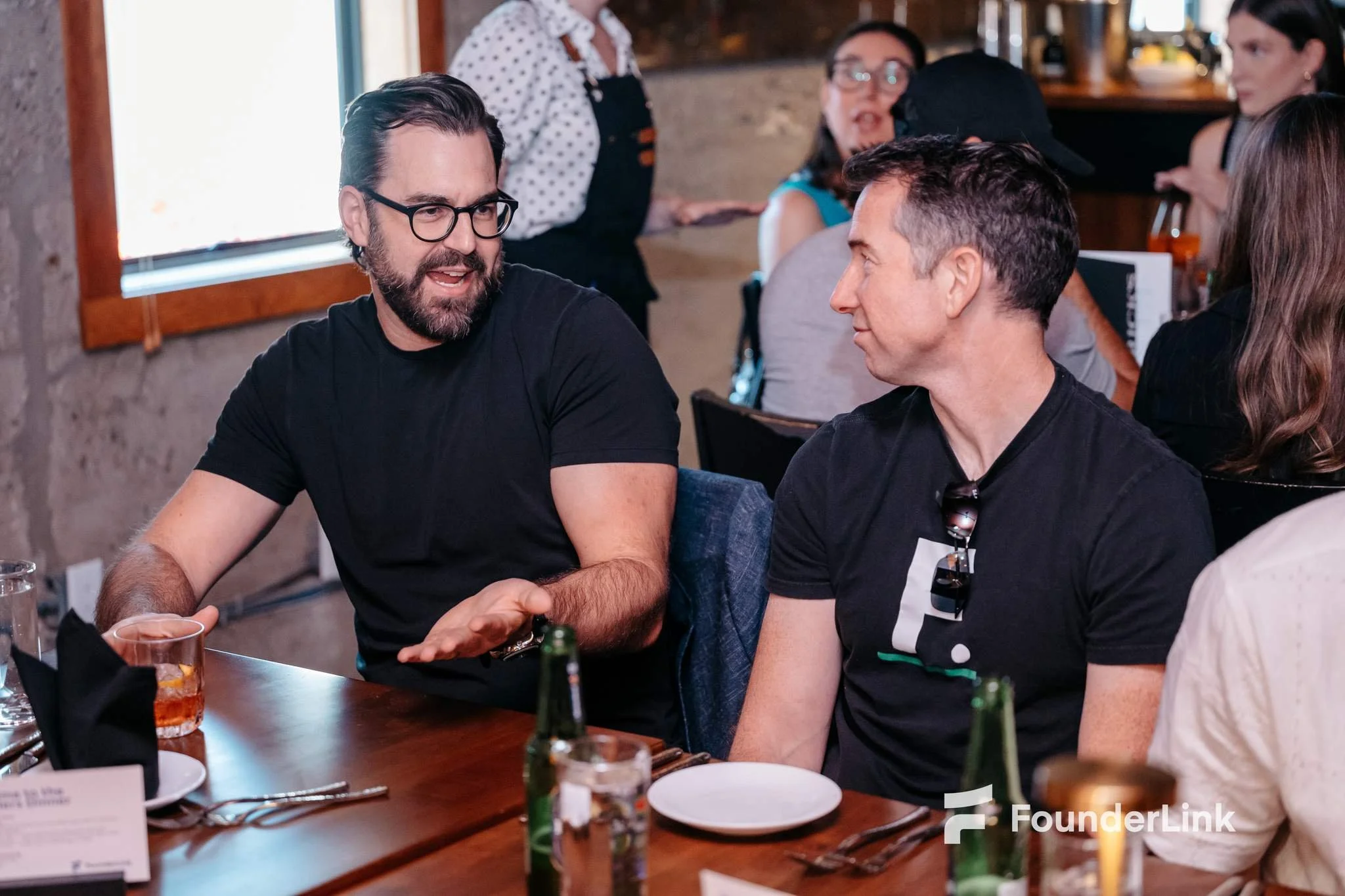 Two men sitting at a table, engaged in conversation, with drinks and plates in front of them, in a restaurant setting with other people in the background, FounderLink event.