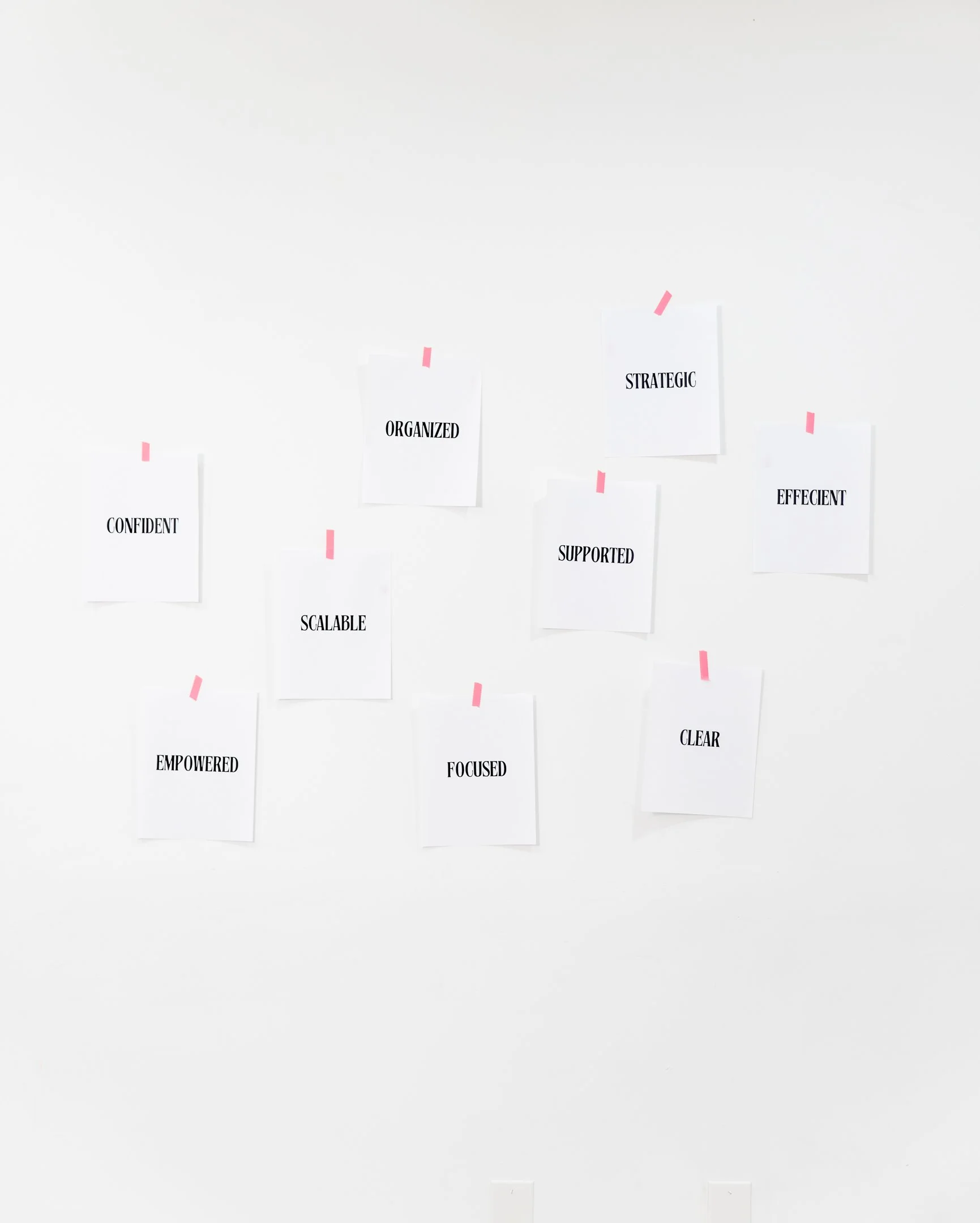 Several pieces of white paper taped on a white wall with pink tape, each with bold black words such as 'CONFIDENT,' 'ORGANIZED,' 'SUPPORTED,' 'SCALABLE,' 'EFFECTIVE,' 'EMPOWERED,' 'FOCUSED,' 'CLEAR,' and 'STRATEGIC.'