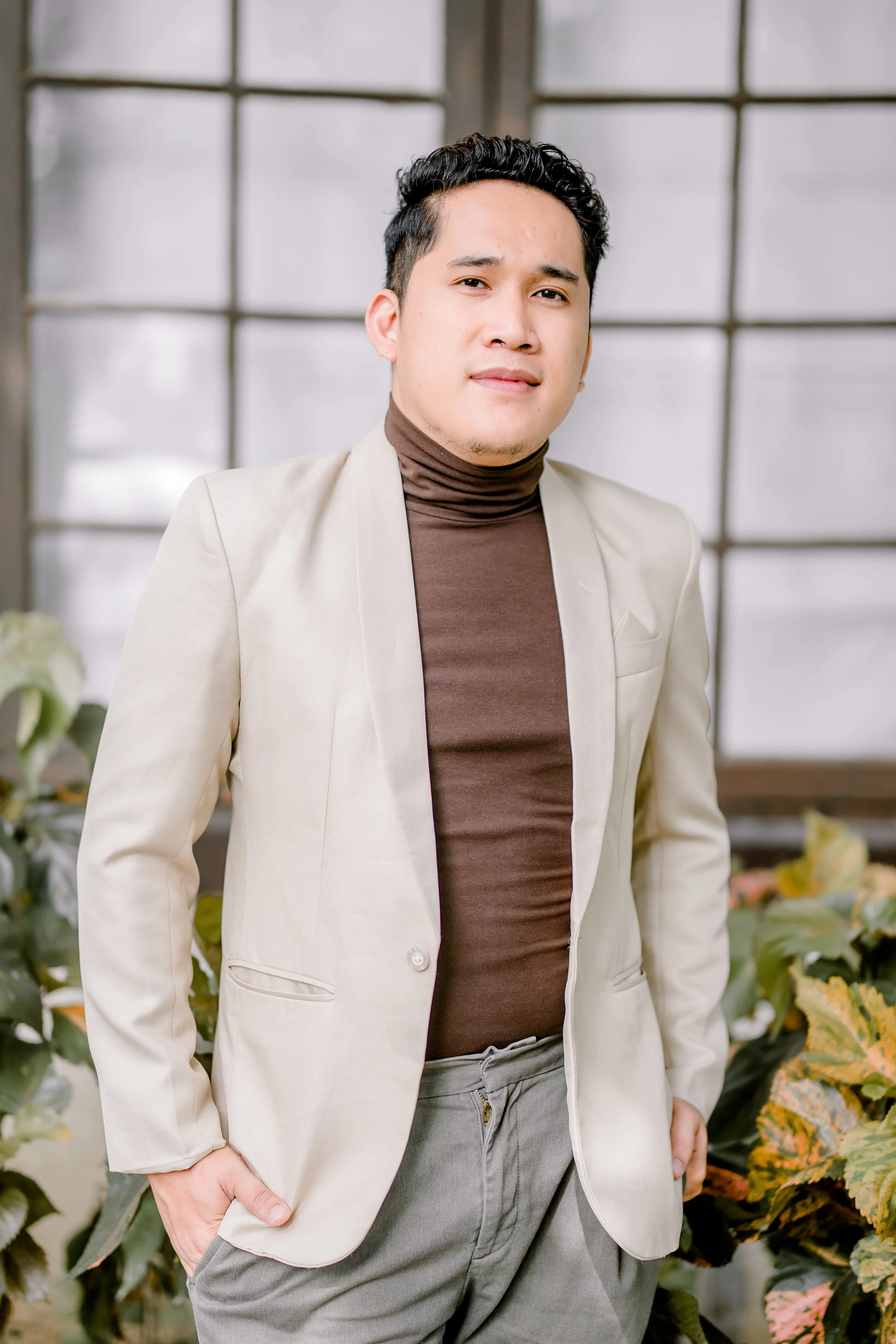 A young man standing indoors in front of large windows with plants around him, wearing a beige blazer, brown turtleneck, and gray pants, with one hand in his pocket.
