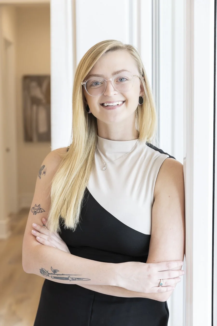 A smiling young woman with blonde hair and glasses standing by a window with her arms crossed, wearing a black and white sleeveless top and earrings.