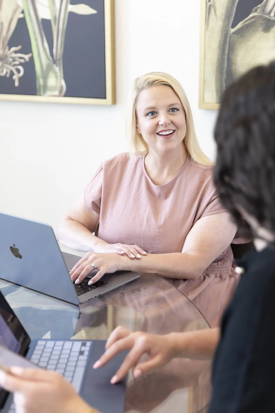 Two women having a discussion at a glass table with laptops in a modern office or meeting room.