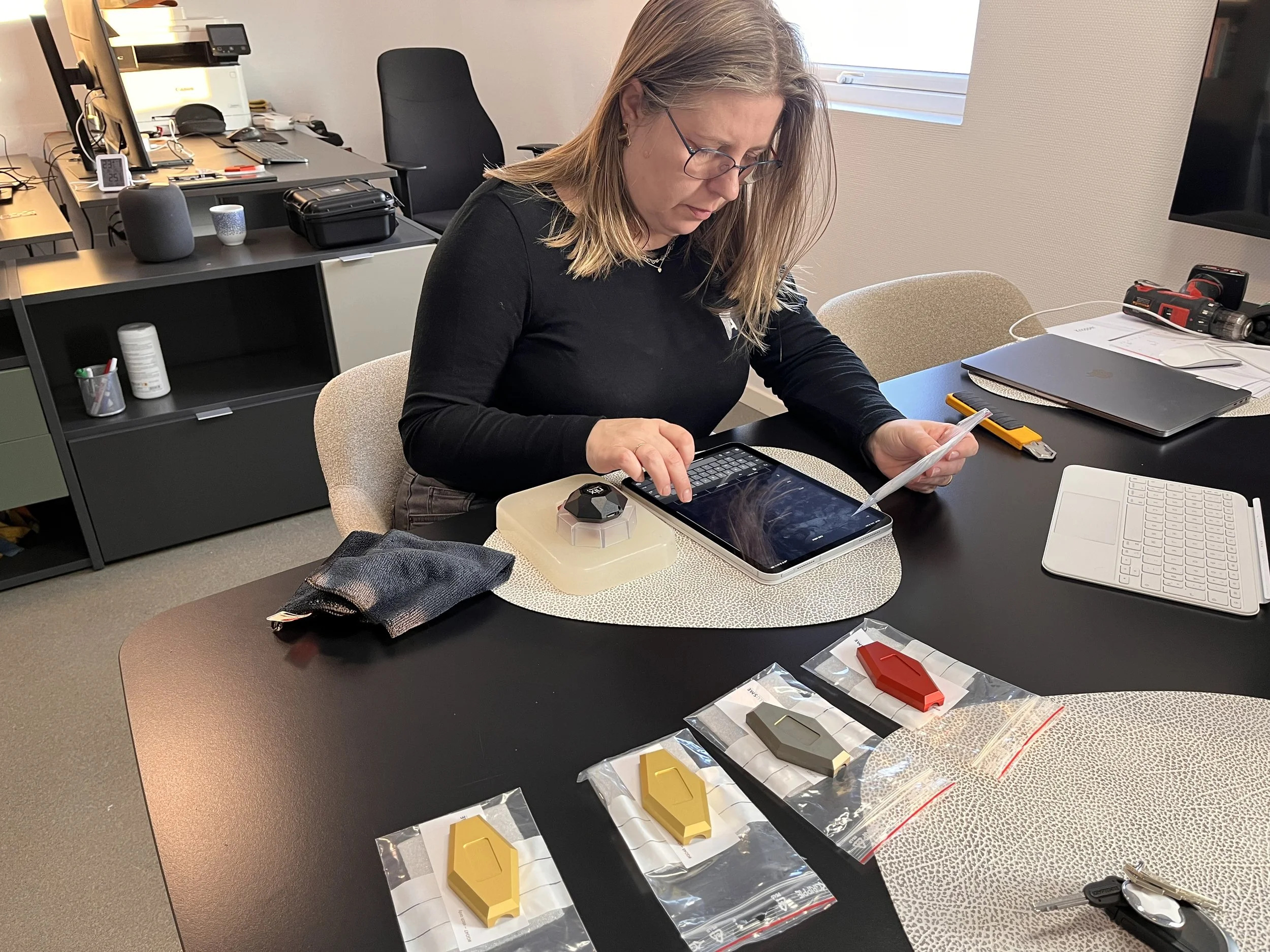 Woman sitting at a table in an office, examining a color sample while surrounded by tools and electronic devices.