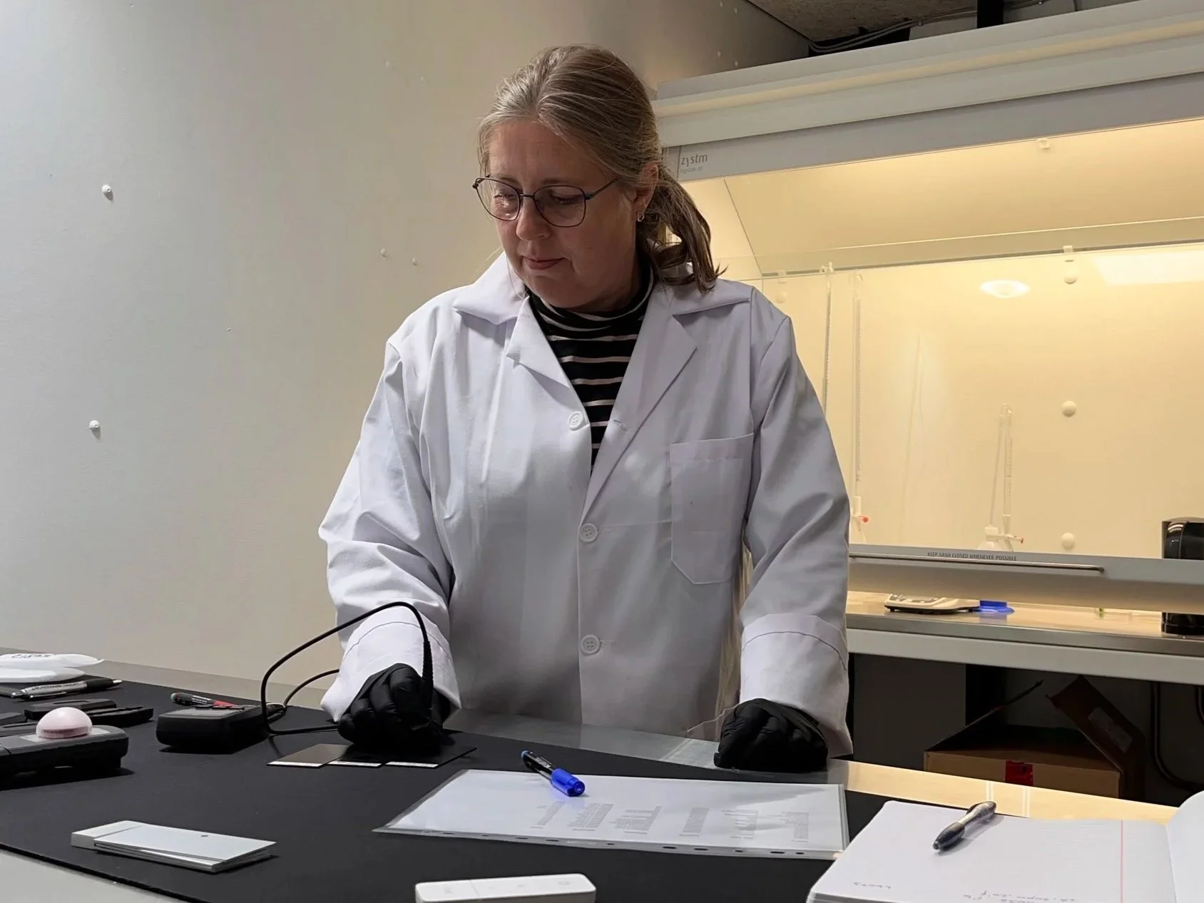 A female scientist in a white lab coat working at a lab bench with various tools and testing equipment.