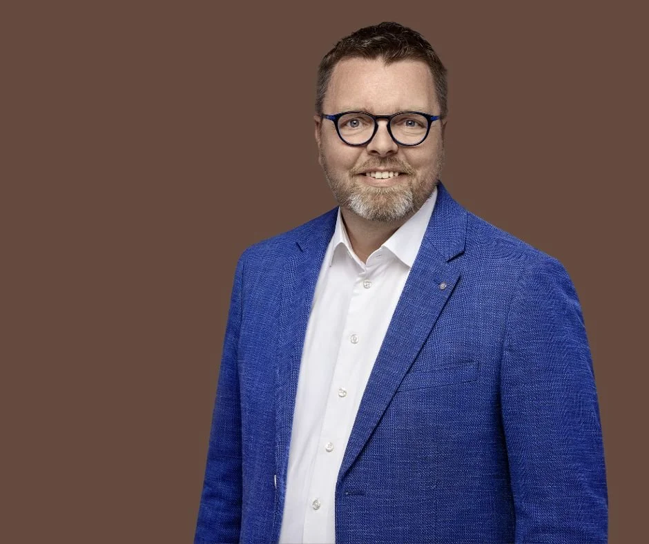 A man with glasses, a beard, and short brown hair, wearing a blue blazer and a white shirt, smiling at the camera against a brown background.