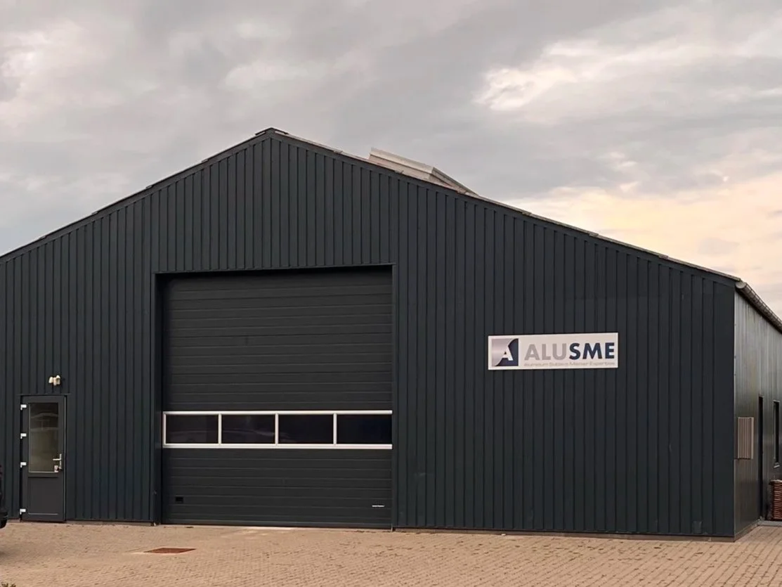 A black metal industrial building with a large garage door and a sign that reads 'ALUSME' the building is headquarter og the company AluSME, located I the municipality og Struer in Denmark.