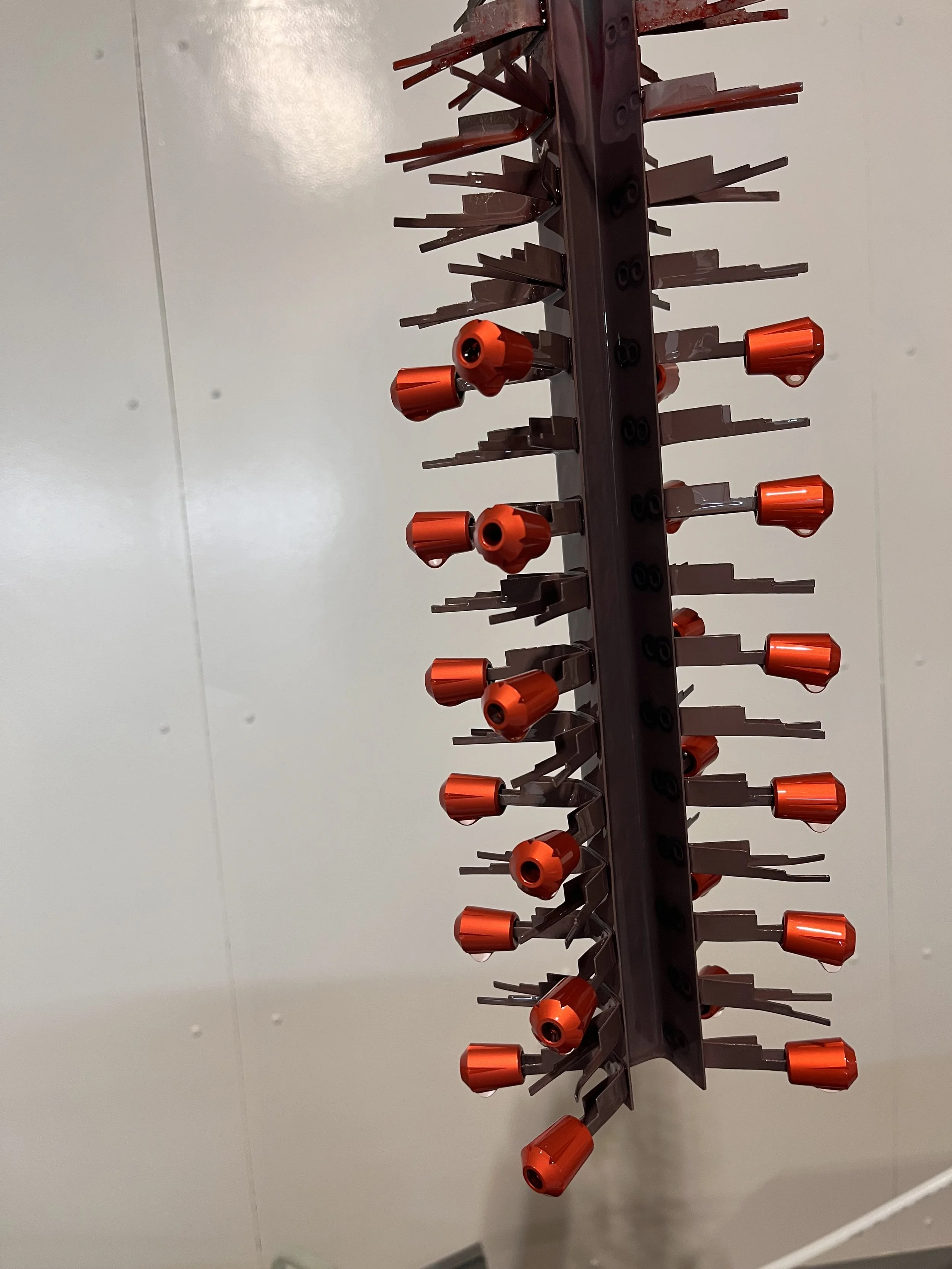 A vertical anodizing rack with multiple red caps attached, positioned against a plain white wall.