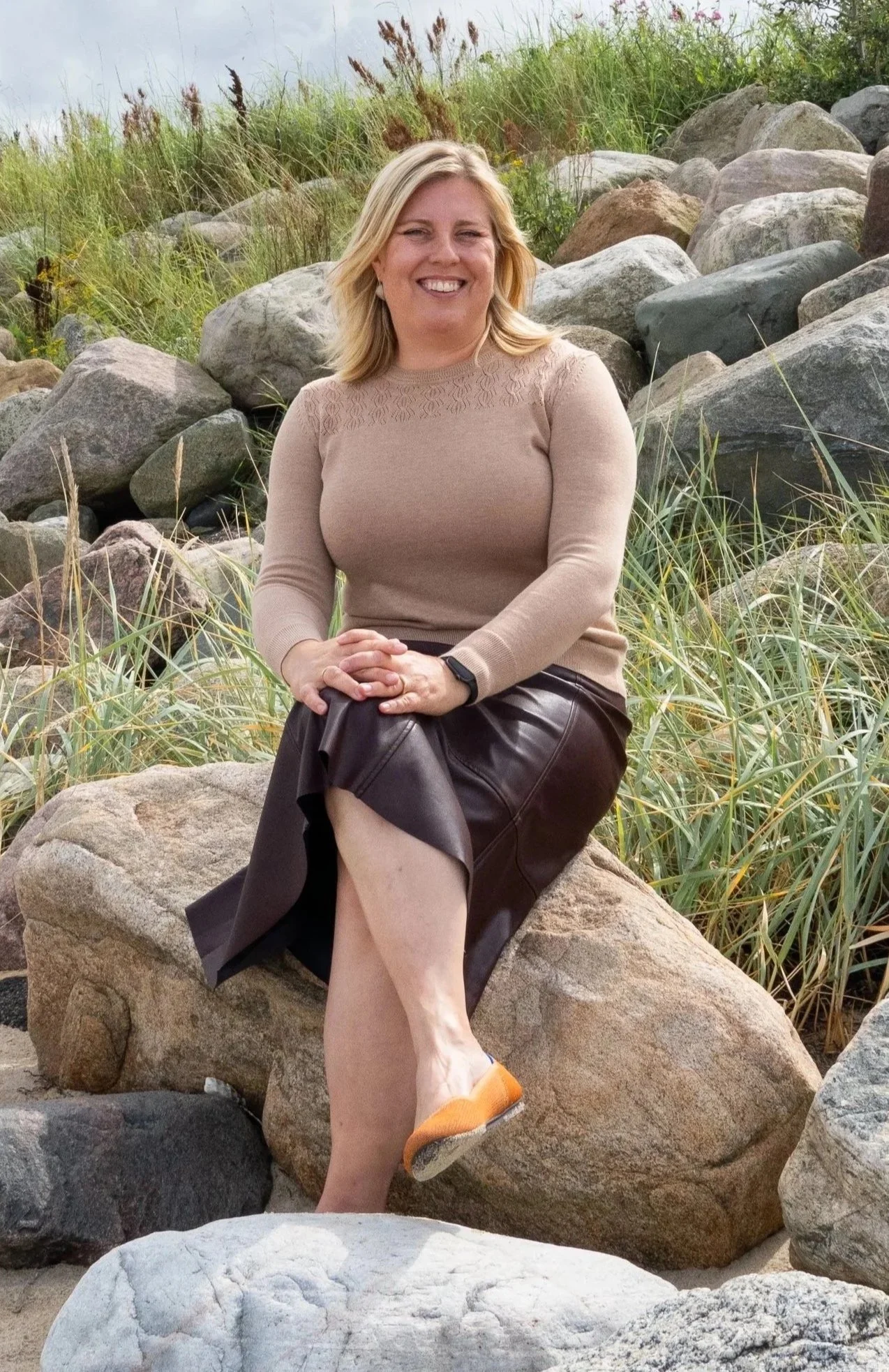 A woman with blonde hair sitting on a large rock among grass and other rocks outdoors, smiling at the camera.