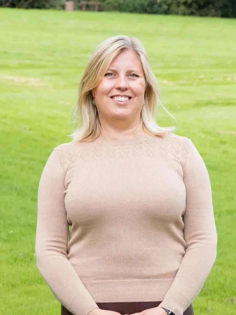 A woman with blonde hair standing outdoors on a grassy area, smiling at the camera.