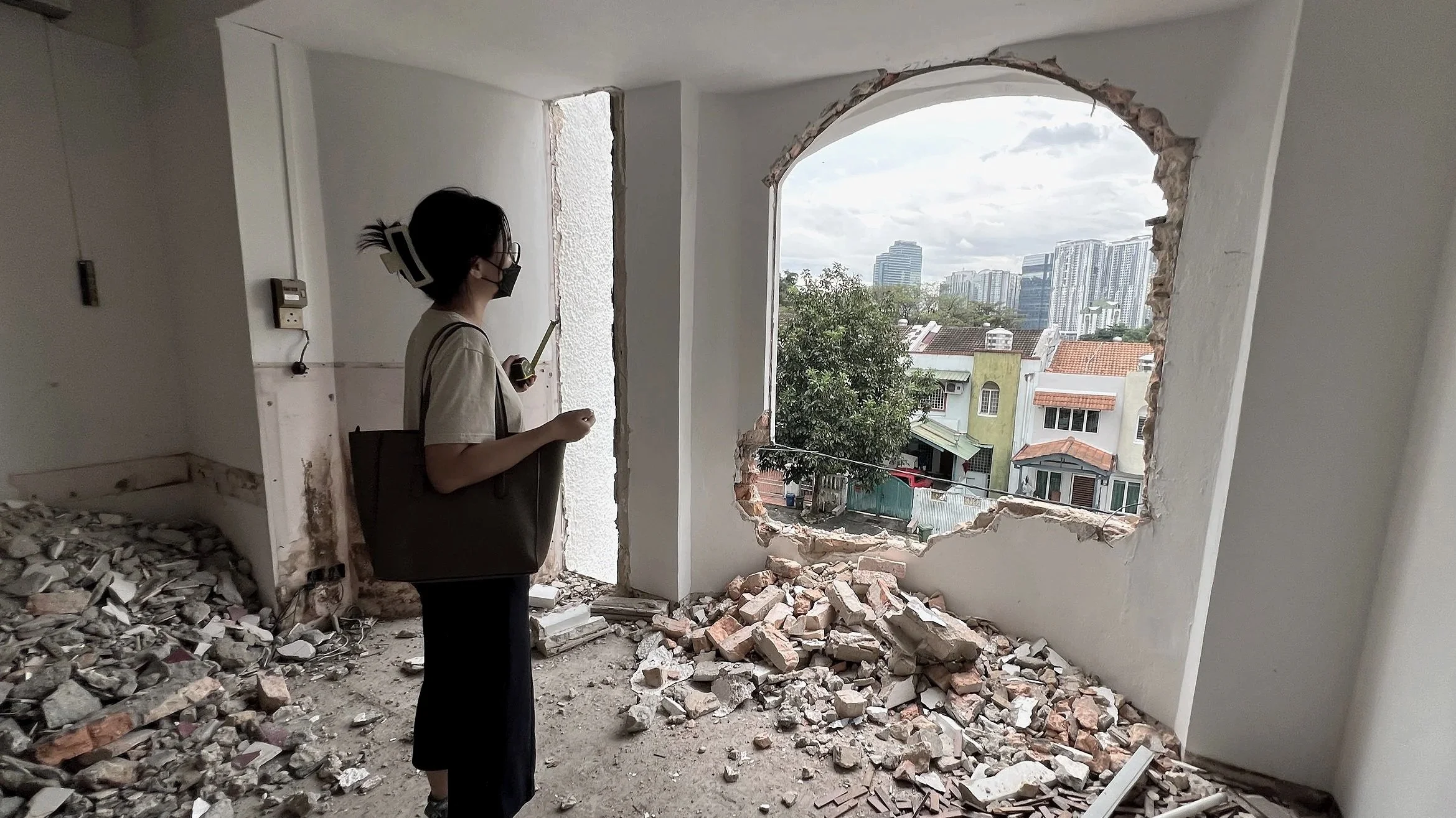 A woman wearing a mask and glasses stands inside a partially demolished room with debris on the floor and a large hole in the wall revealing a cityscape with buildings and trees outside.