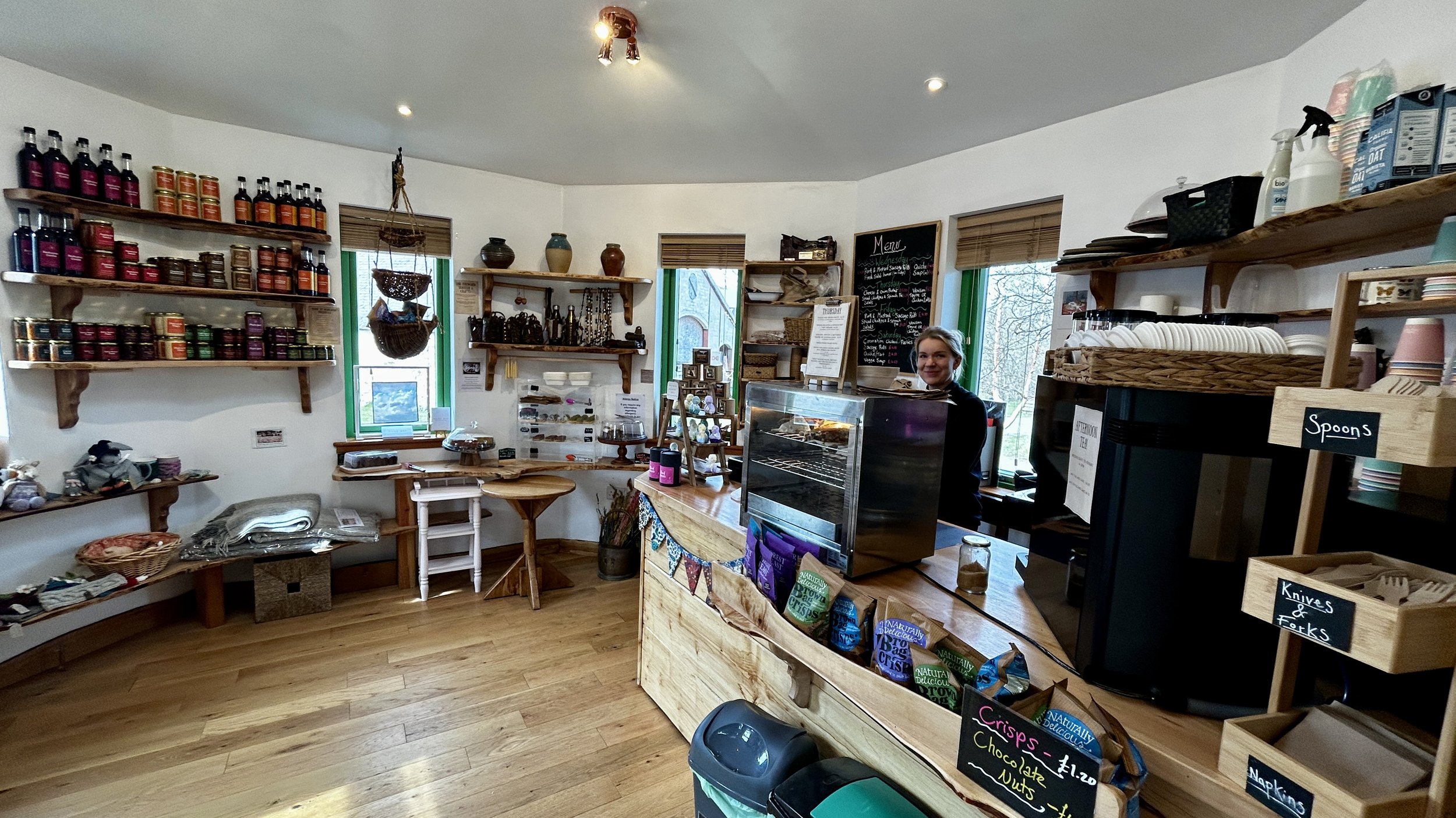 Interior of a cozy shop with wooden shelves holding muffins, tea canisters, and decorative items, a counter with snacks and chocolates, and a smiling woman behind the counter.