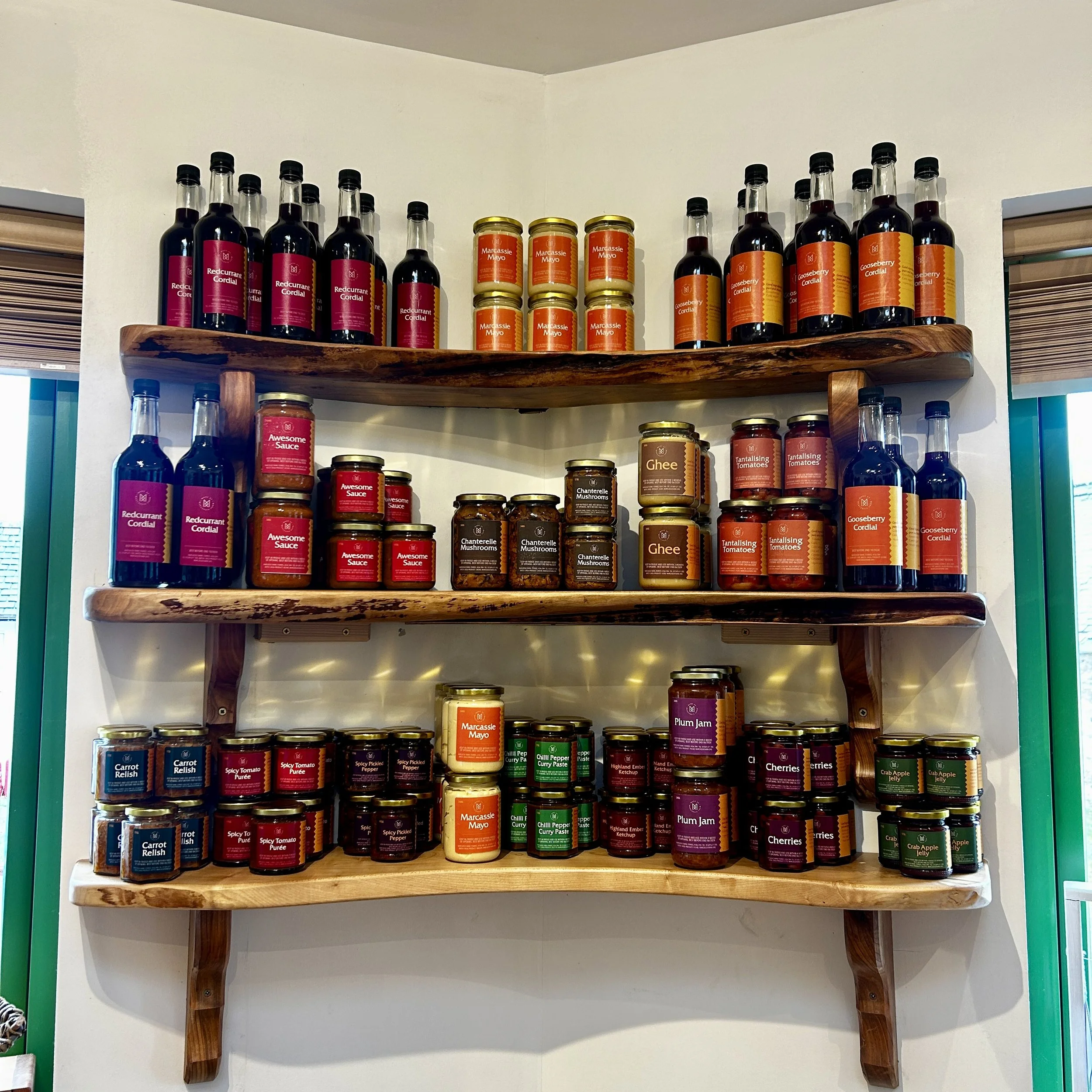 Wooden shelves filled with jars and bottles of various condiments and sauces, including cherry jam, ghee, curry paste, and tomato relish.