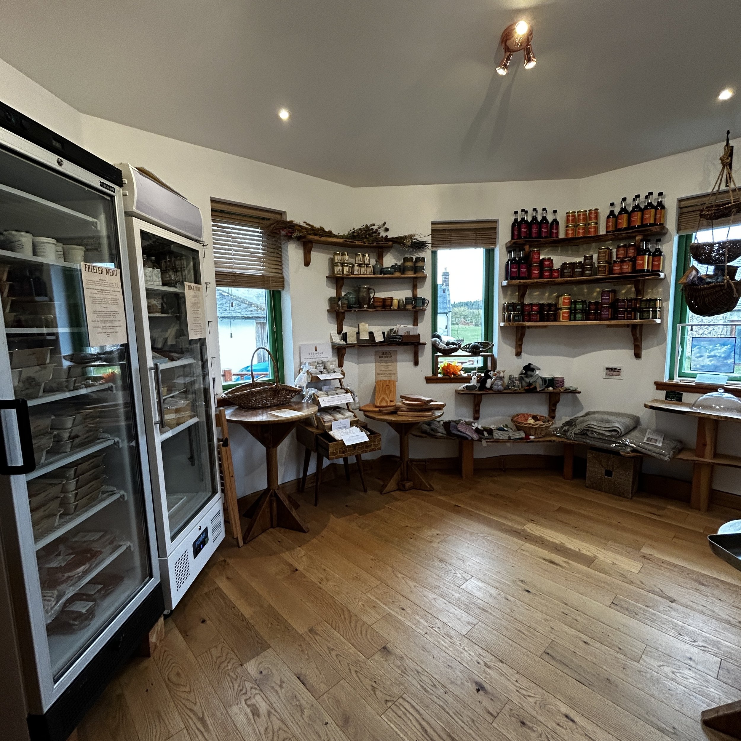 Inside a small shop with wooden floors, displaying jars and bottles on shelves, along with various home goods and textiles on tables and benches, and a freezer with glass doors on the left.