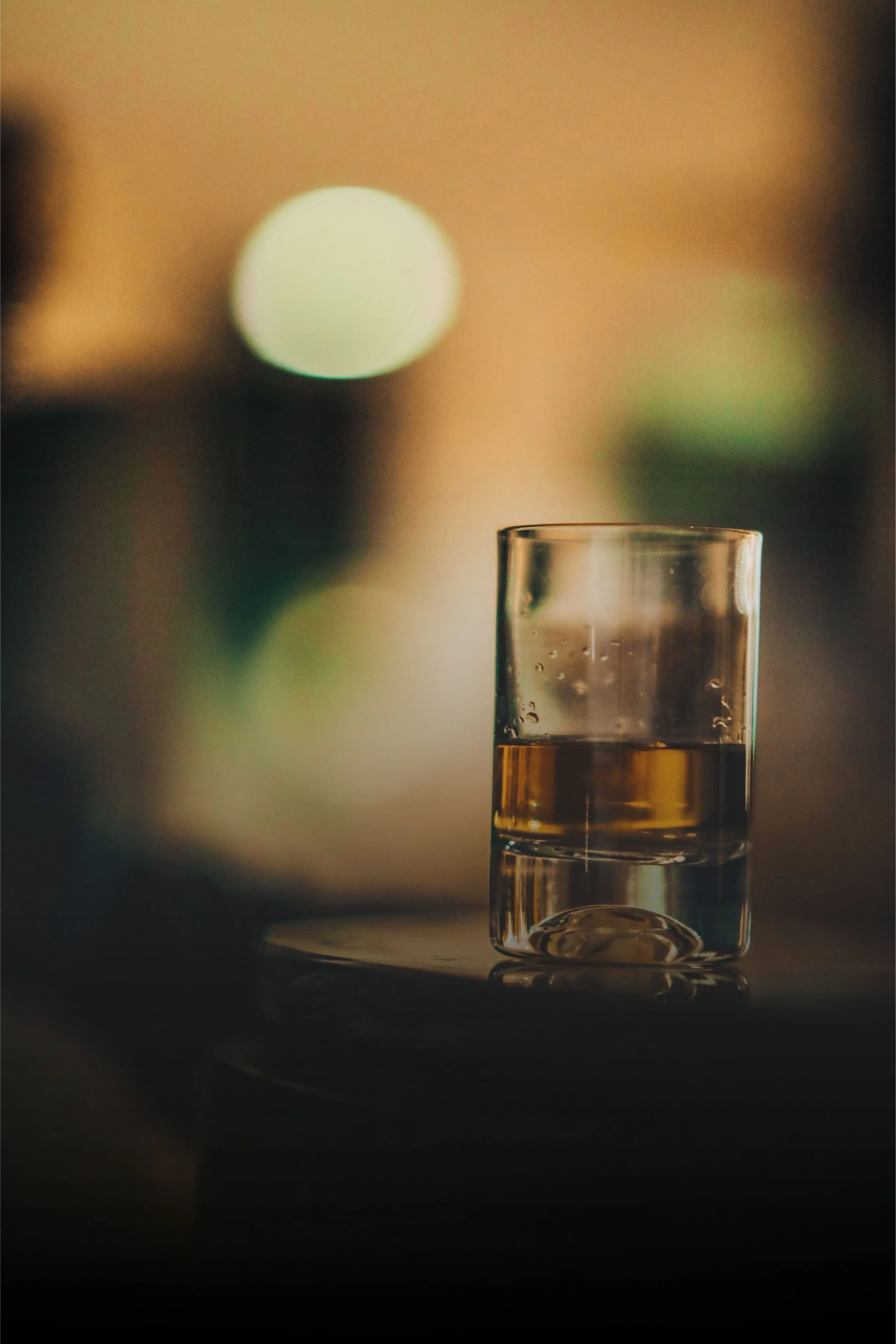 A clear glass of whiskey placed on a dark surface, with a blurry background of warm lights.