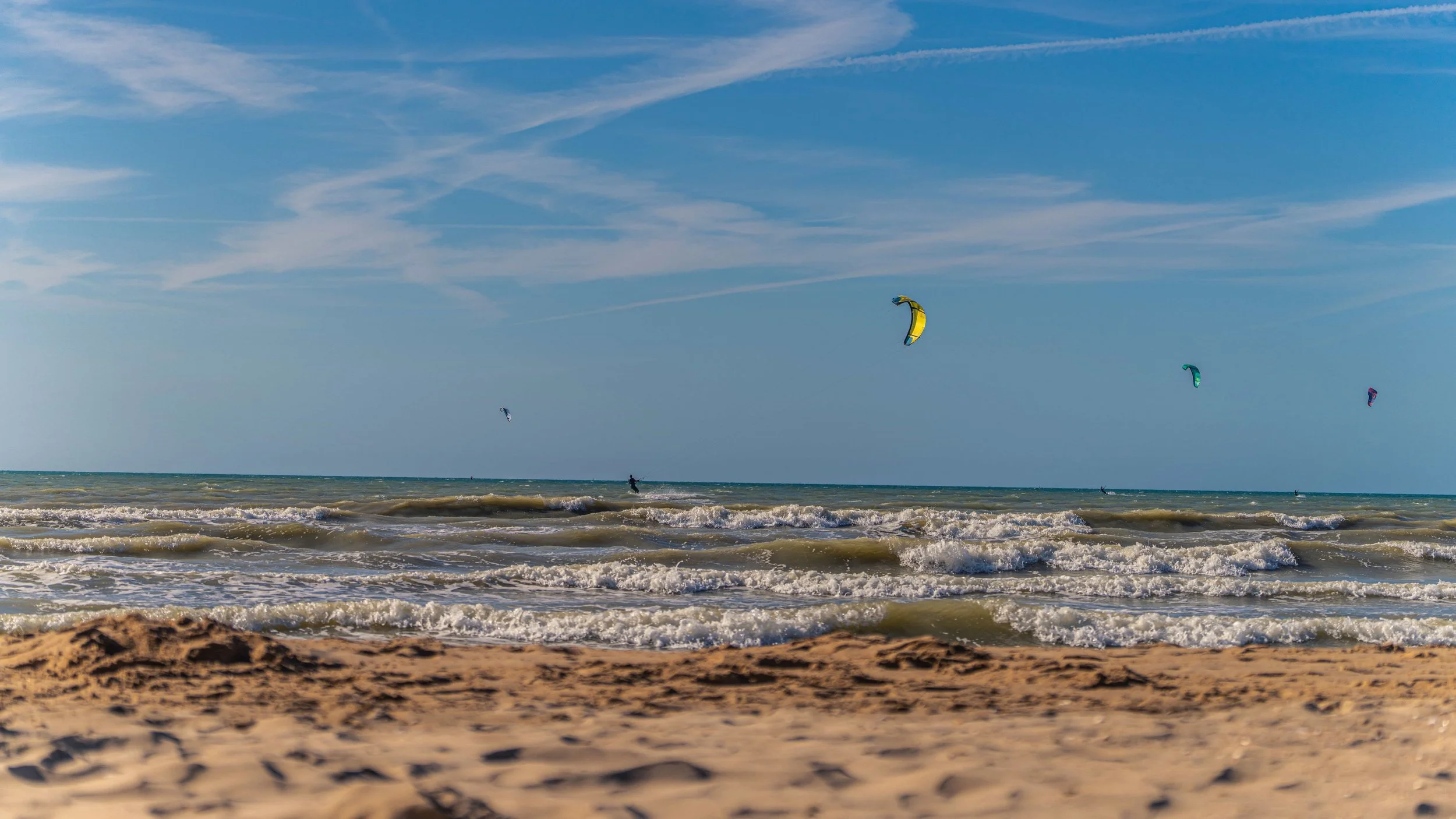 Kite surfers riding waves at the beach under a blue sky with some clouds.