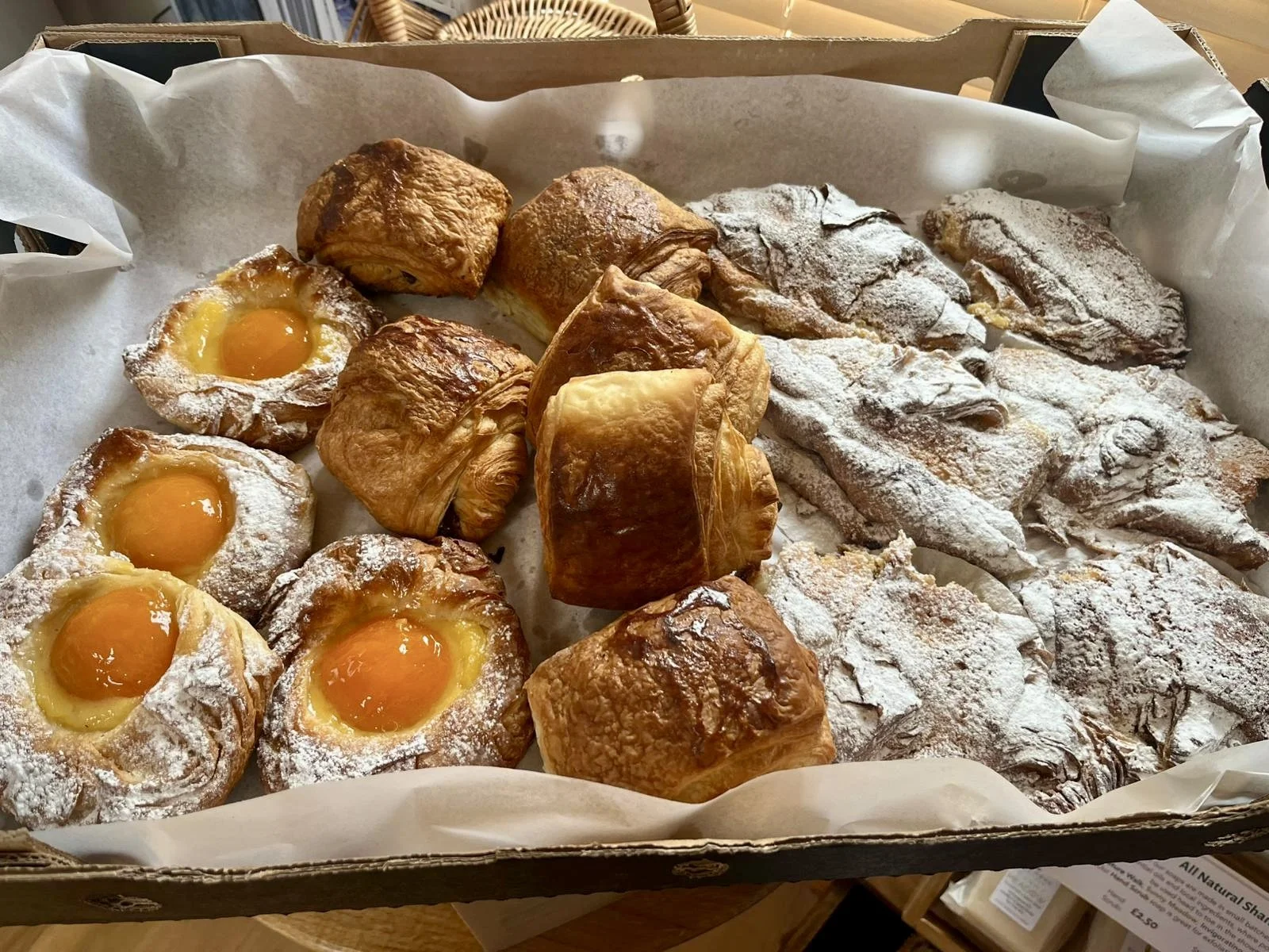 Assorted baked goods, including custard-filled pastries, croissants, and powdered sugar-dusted pastries, inside a cardboard box lined with parchment paper.