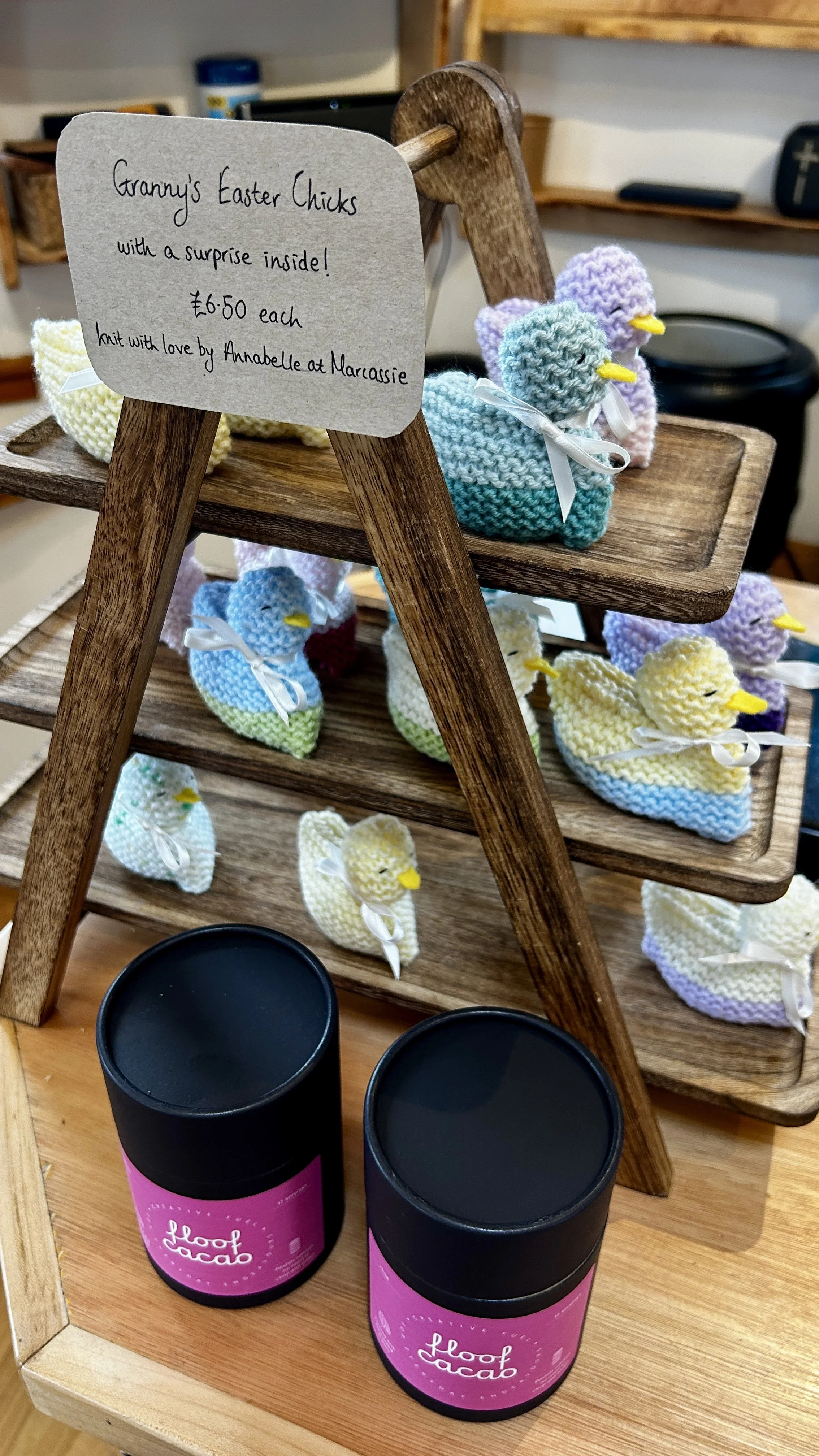 A display of colorful knitted chick toys on a wooden three-tiered stand, with a sign describing them as Granny's Easter chicks with a surprise inside, priced at £6.50 each, handmade by Annabelle at Marcassie. There are also two jars of food cacao in 