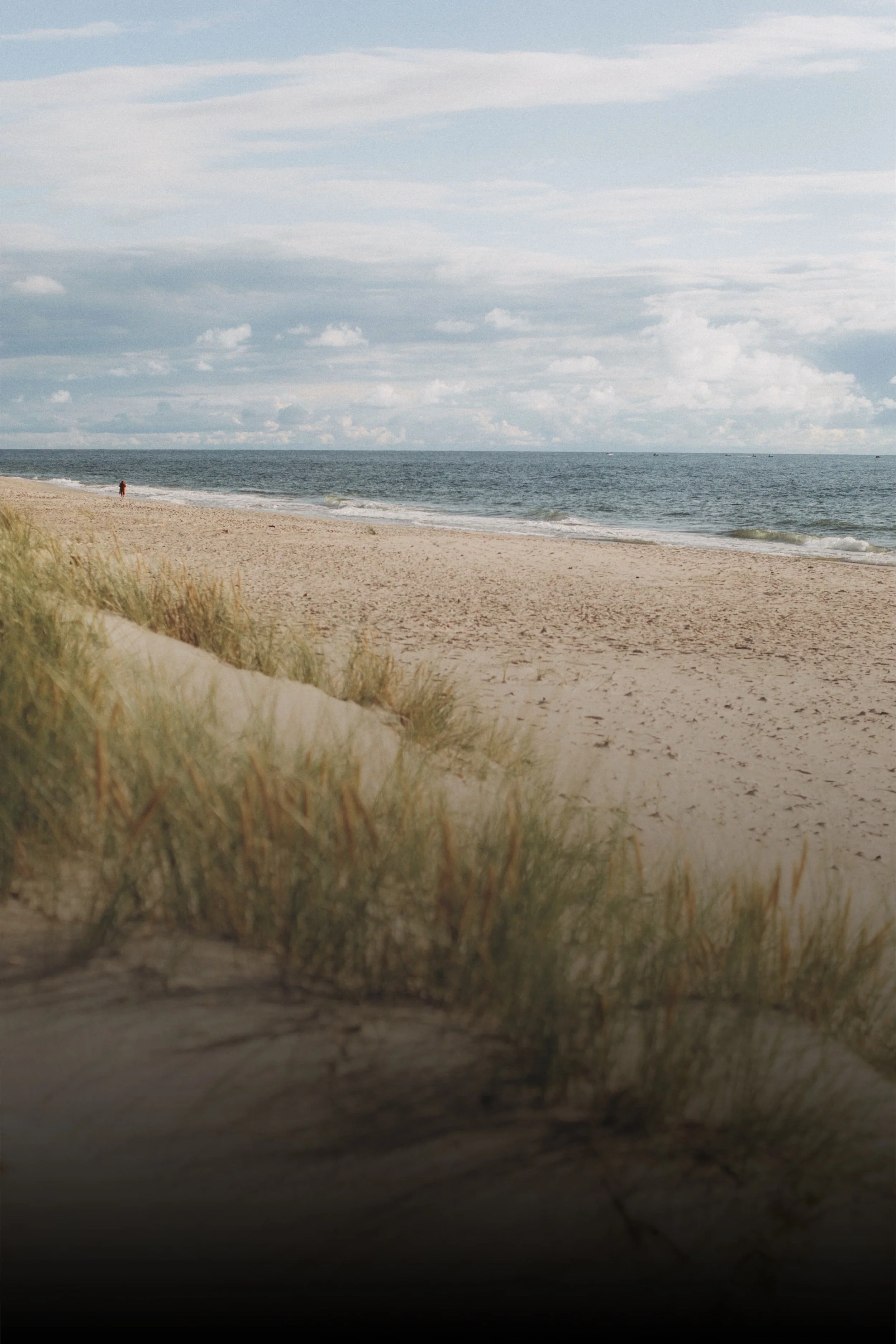 View of a beach with sand dunes and grass in the foreground, and the ocean with small waves and a cloudy sky in the background.
