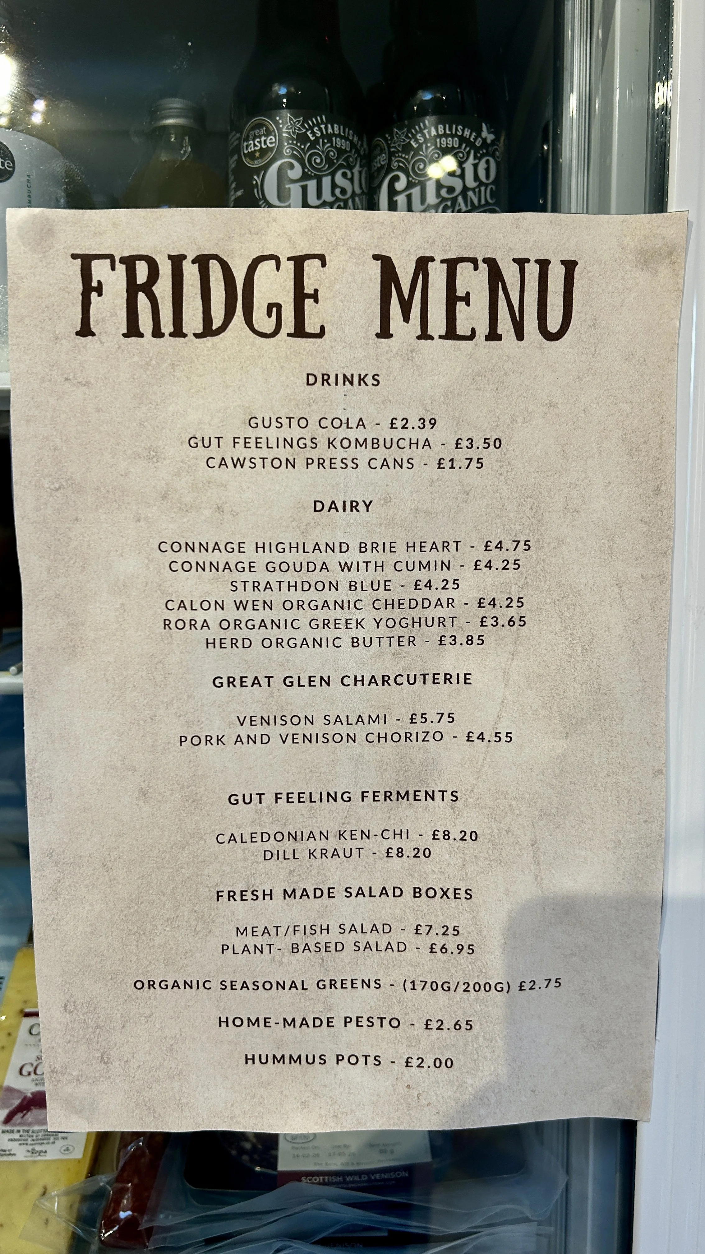 A fridge door displaying a menu with various drinks, dairy products, charcuterie, gut feeling ferments, salad boxes, greens, and dips. Items include Gusto Cola, kombucha, canned drinks, cheeses, yogurts, salami, chorizo, ferments, salads, greens, pes