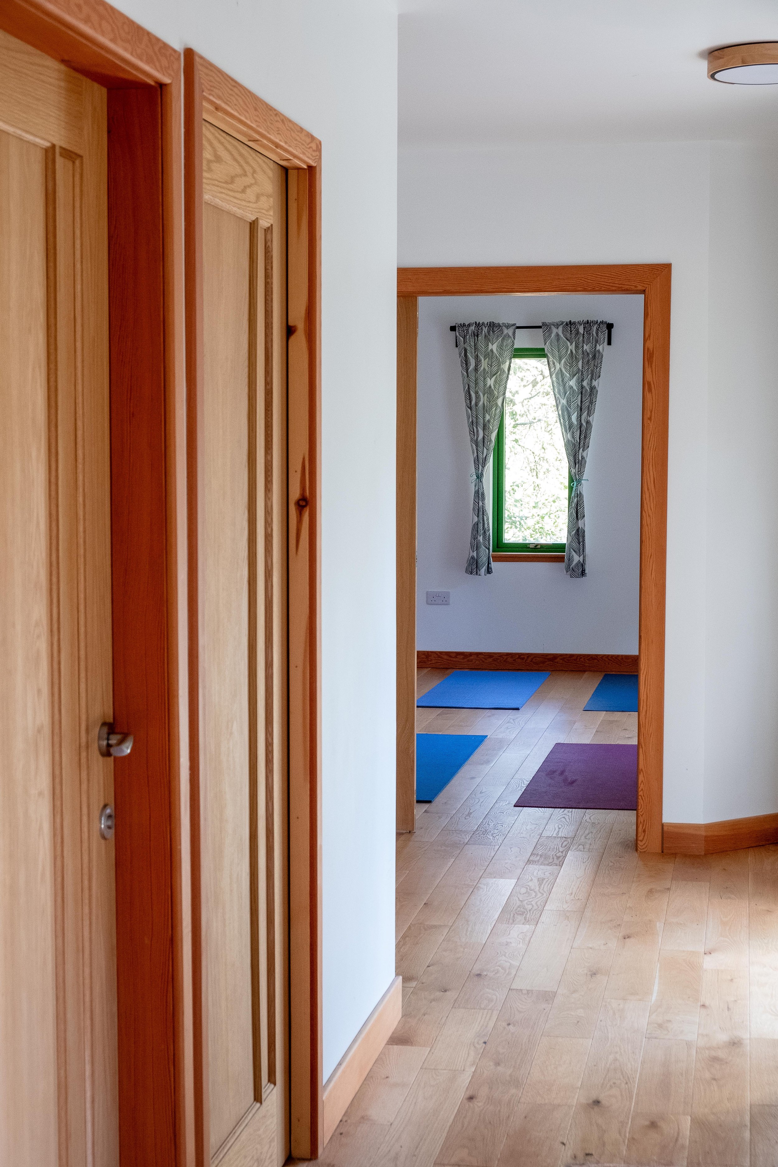 Interior hallway with wooden doors, leading to a room with yoga mats and a window with patterned curtains.