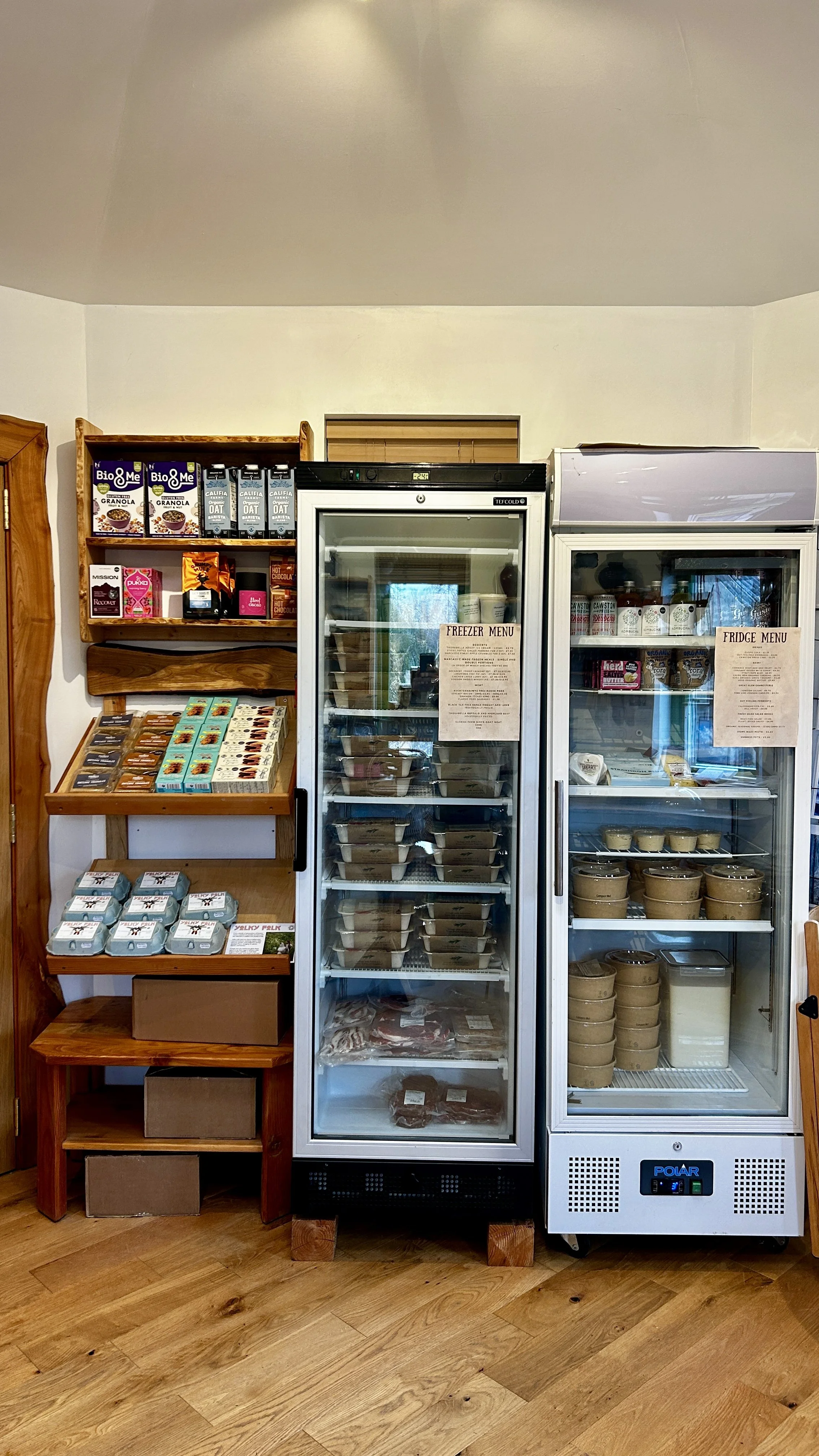 Inside a small grocery or deli store, with two glass-door refrigerators filled with frozen and refrigerated foods, shelves with boxed items, and a wooden shelf displaying various snacks and packaged goods.