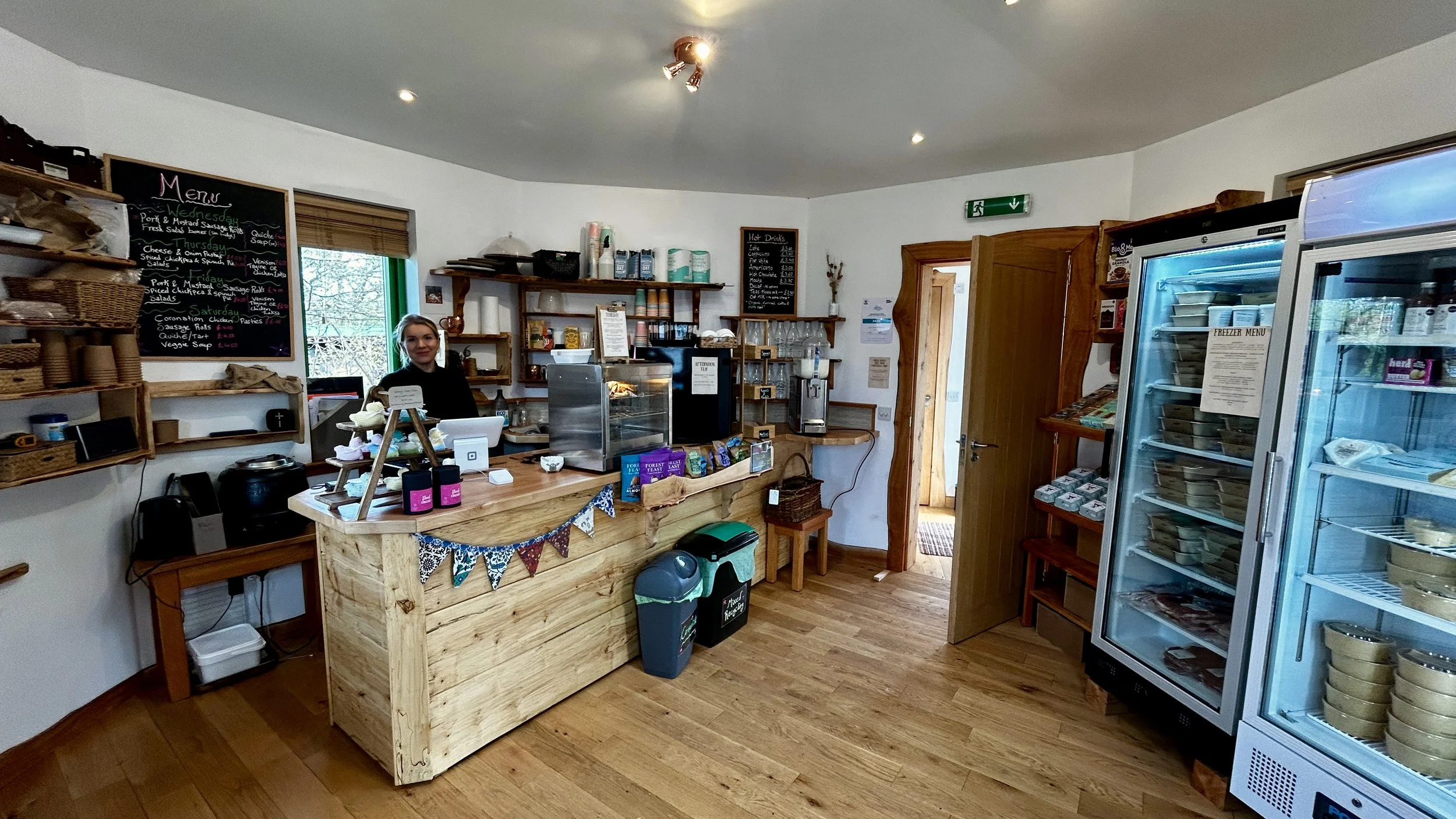 Inside a cozy cafe with a wooden counter, shelves with supplies, a woman behind the counter smiling, and refrigerated display cases with food items.