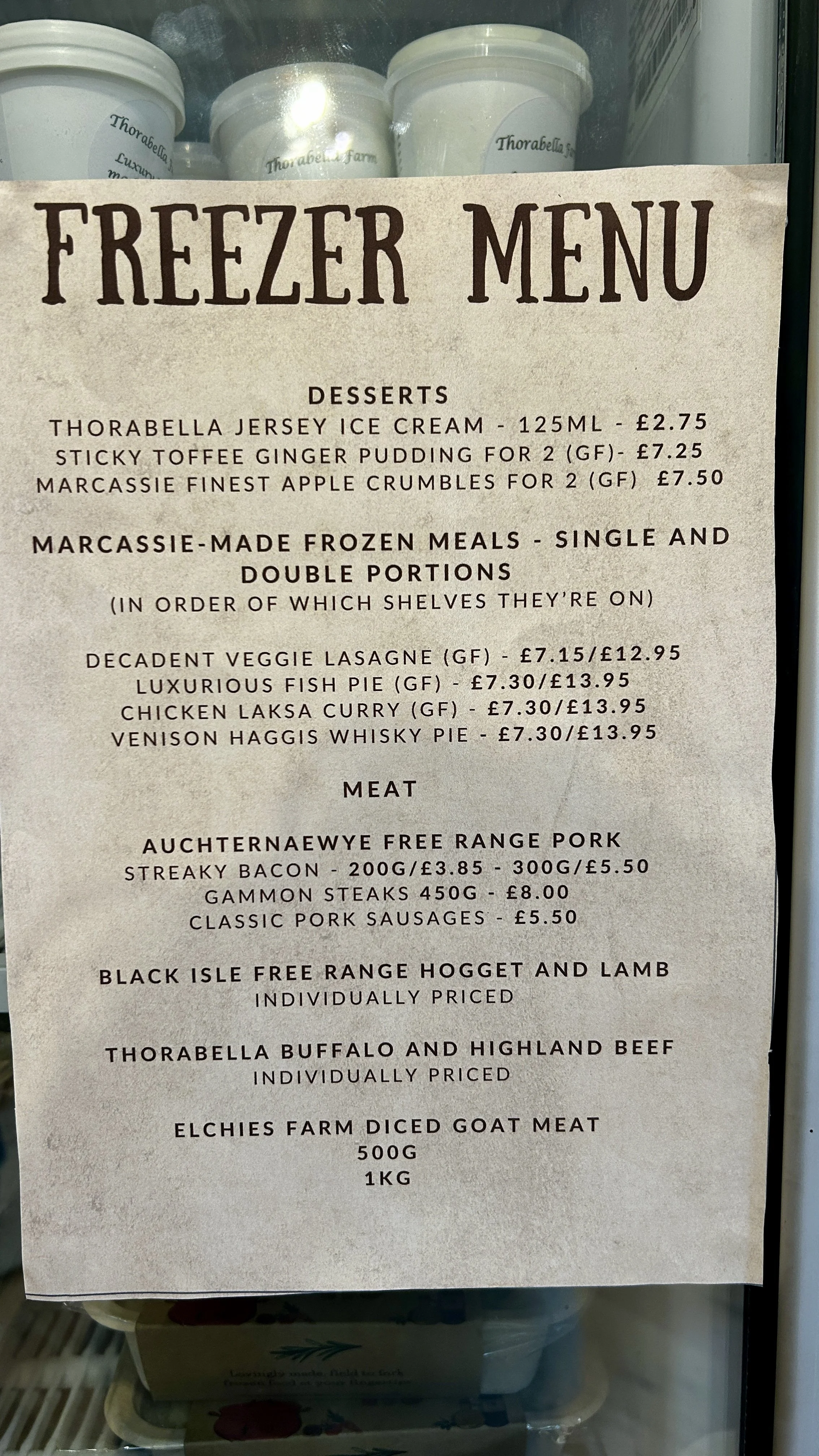 Freezer menu sign with desserts, frozen meals, and meats including ice cream, puddings, lasagne, fish pie, curry, venison, pork, lamb, beef, and goat meat.