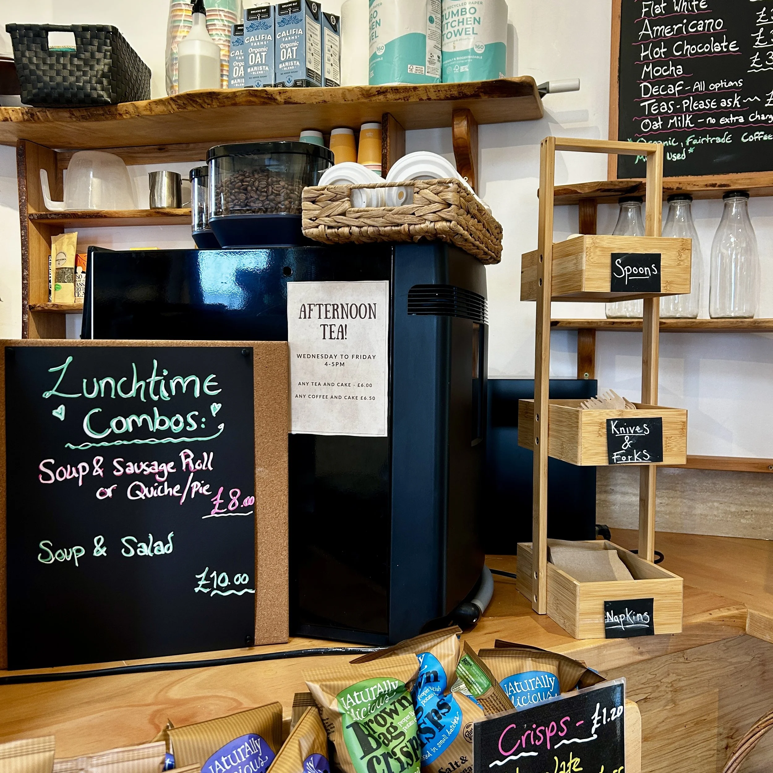 Coffee shop counter with black coffee machine, bowls, baskets, and a wooden shelf for utensils. Chalkboards display menu and prices, with snacks placed in front.