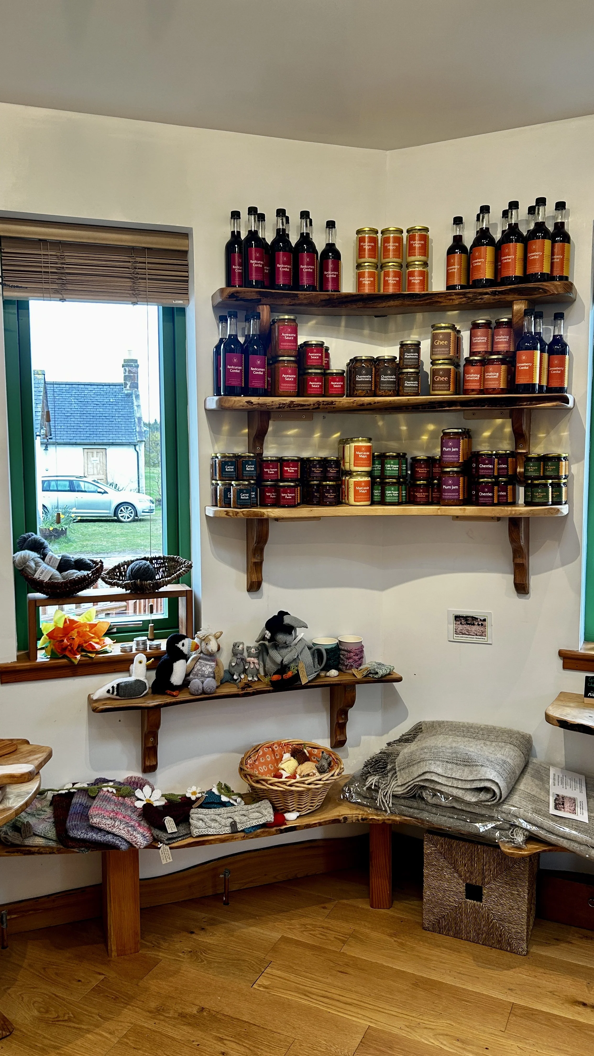 Shelves displaying jams, preserves, and condiments; knitted toys and accessories on a wooden bench near a window in a cozy shop.