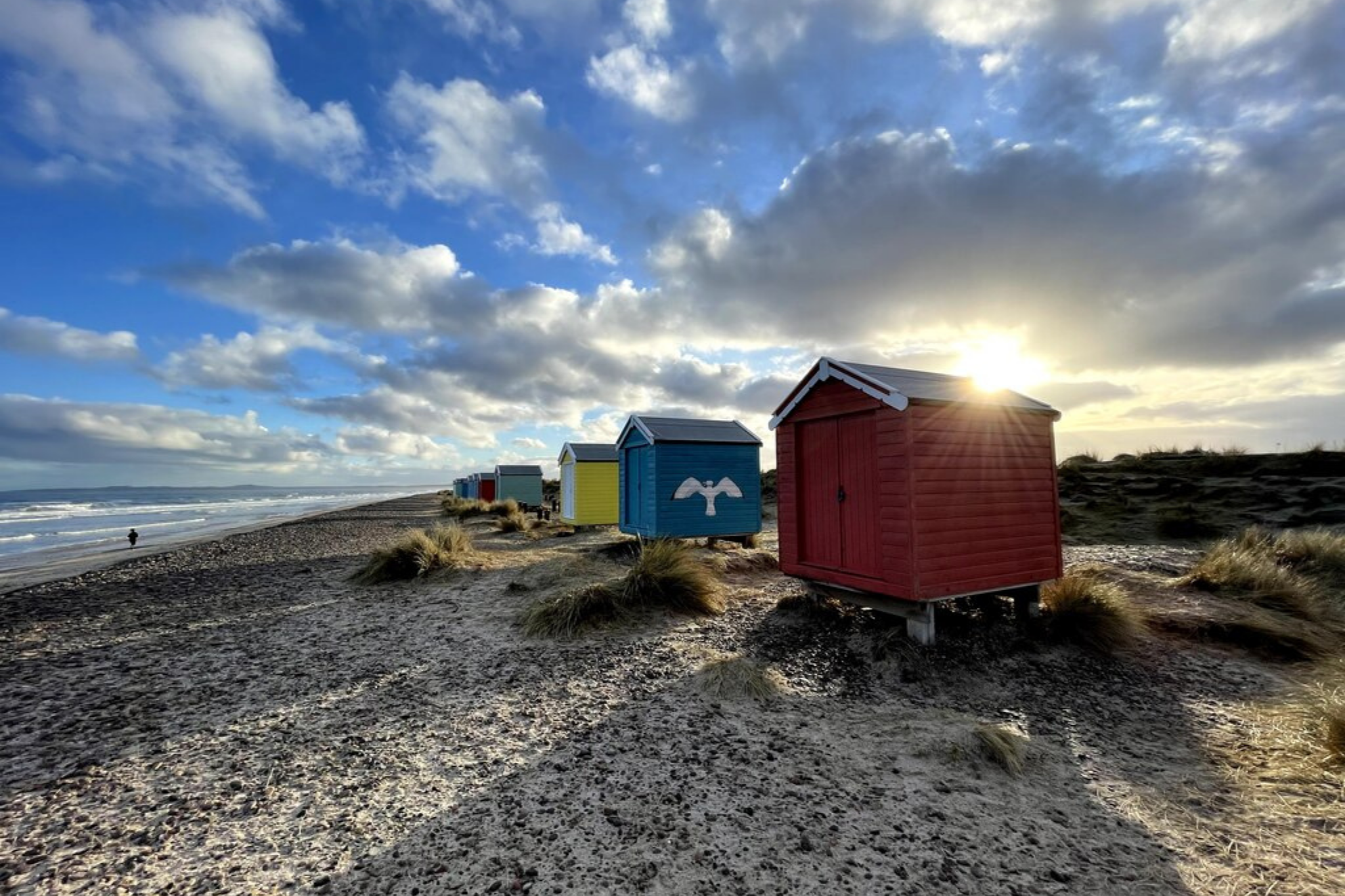 Colorful beach huts along the shoreline at sunset, with the sun partially hidden behind the red hut, a partly cloudy sky, ocean waves, and a person walking along the beach in the distance.