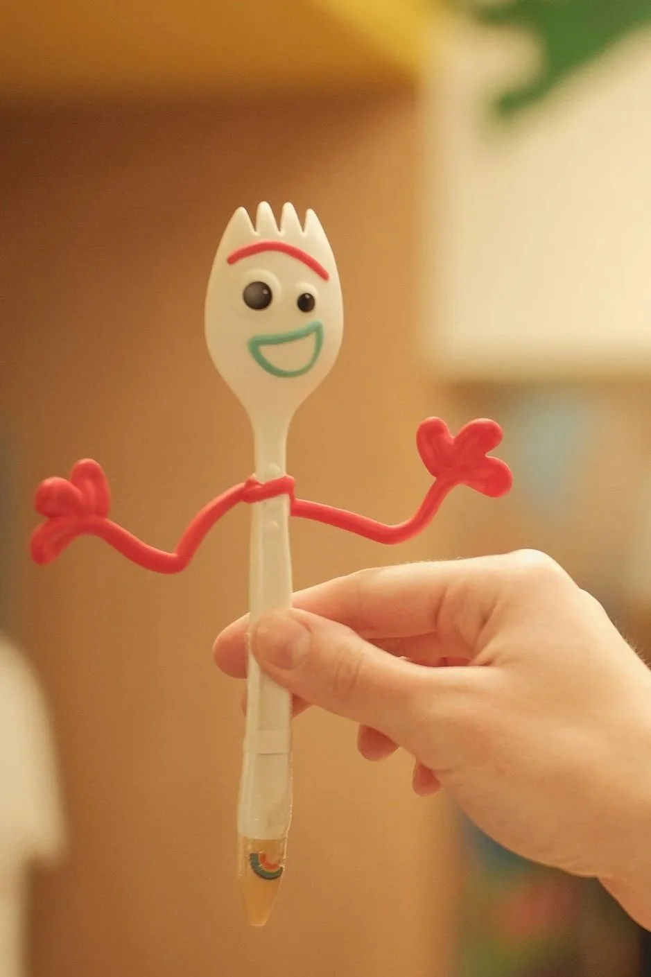 Cucchiaio con faccia sorridente, occhi vivaci e braccia fatte di elastici rossi, tenuto da una mano.
