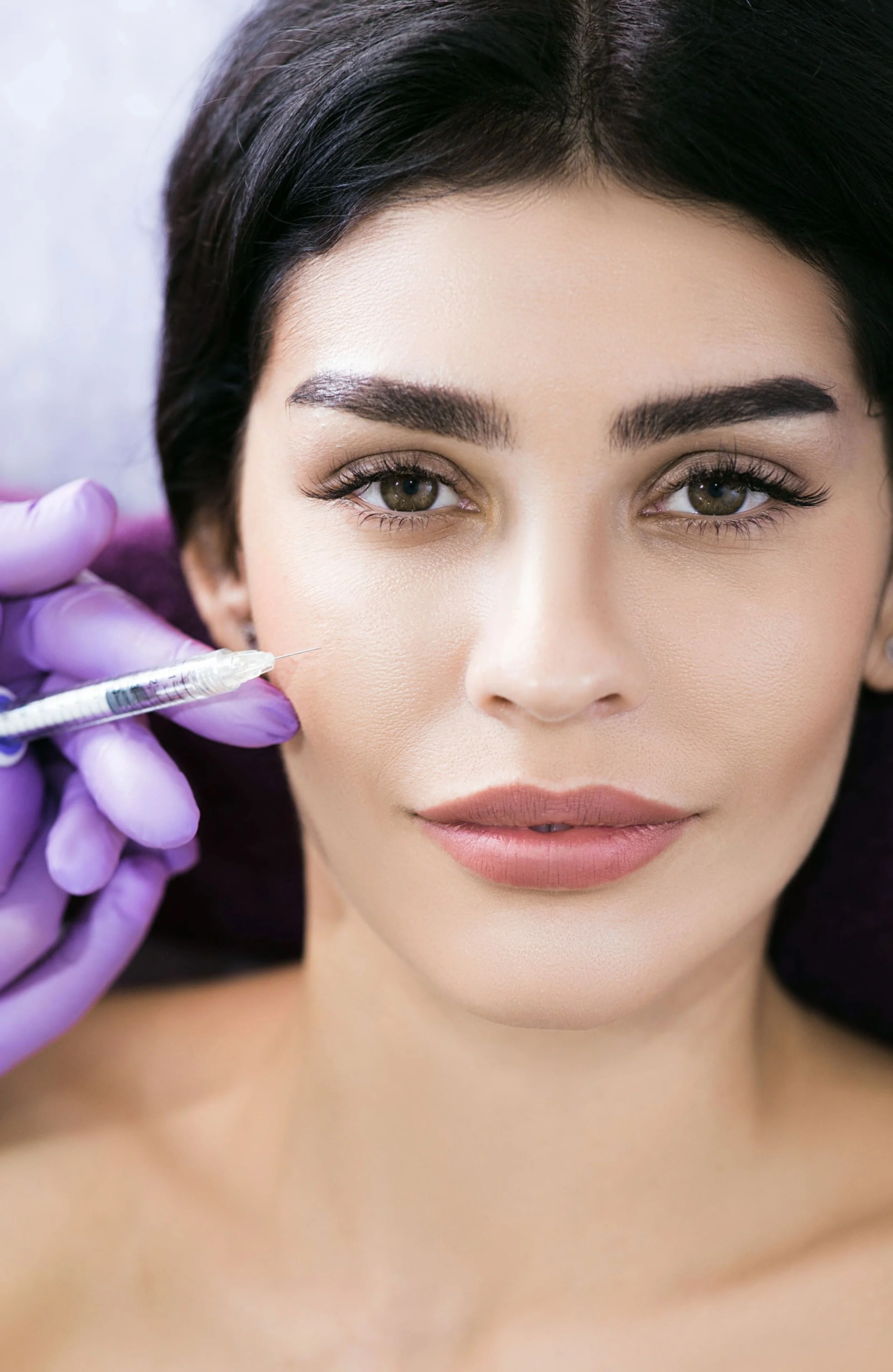 Una donna con pelle liscia e trucco naturale, che riceve un trattamento di medicina estetica con un'ago, mentre è sdraiata su un lettino.