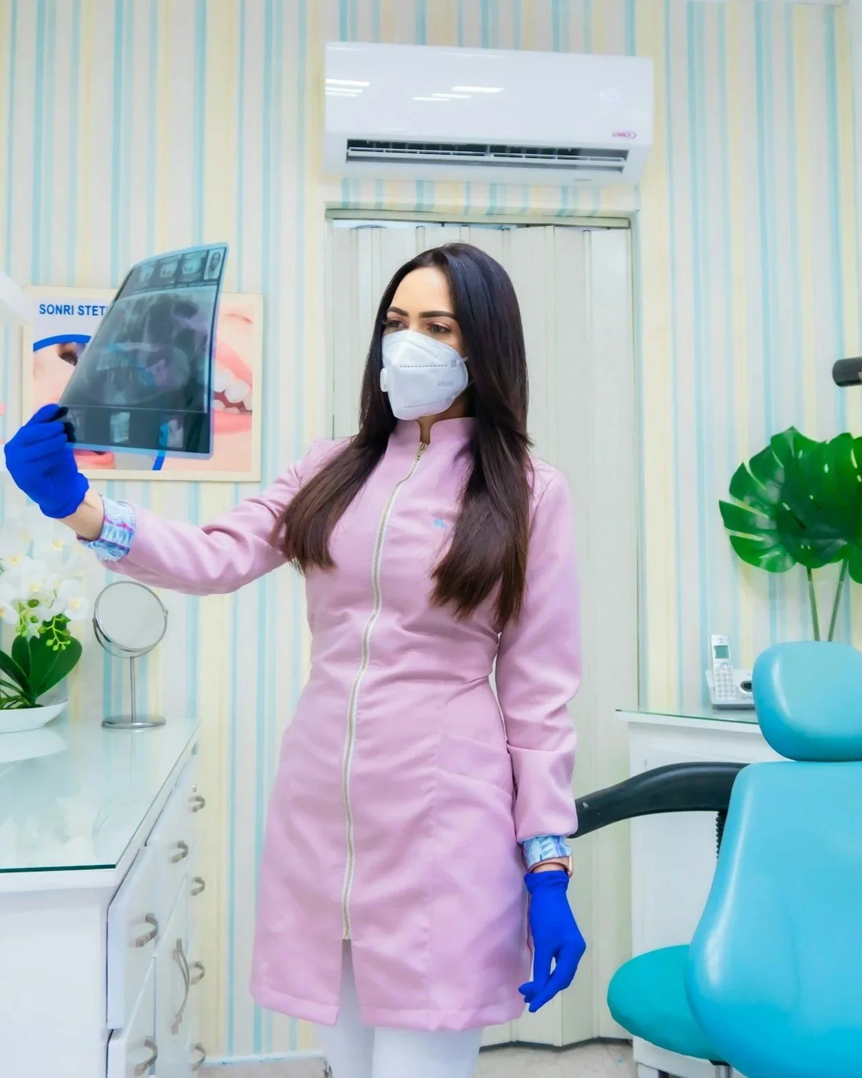 Donna studio dentistico con mascherina, guanti blu, che guarda un radiografia dentale in uno studio con arredi clinici e decorazioni floreali.