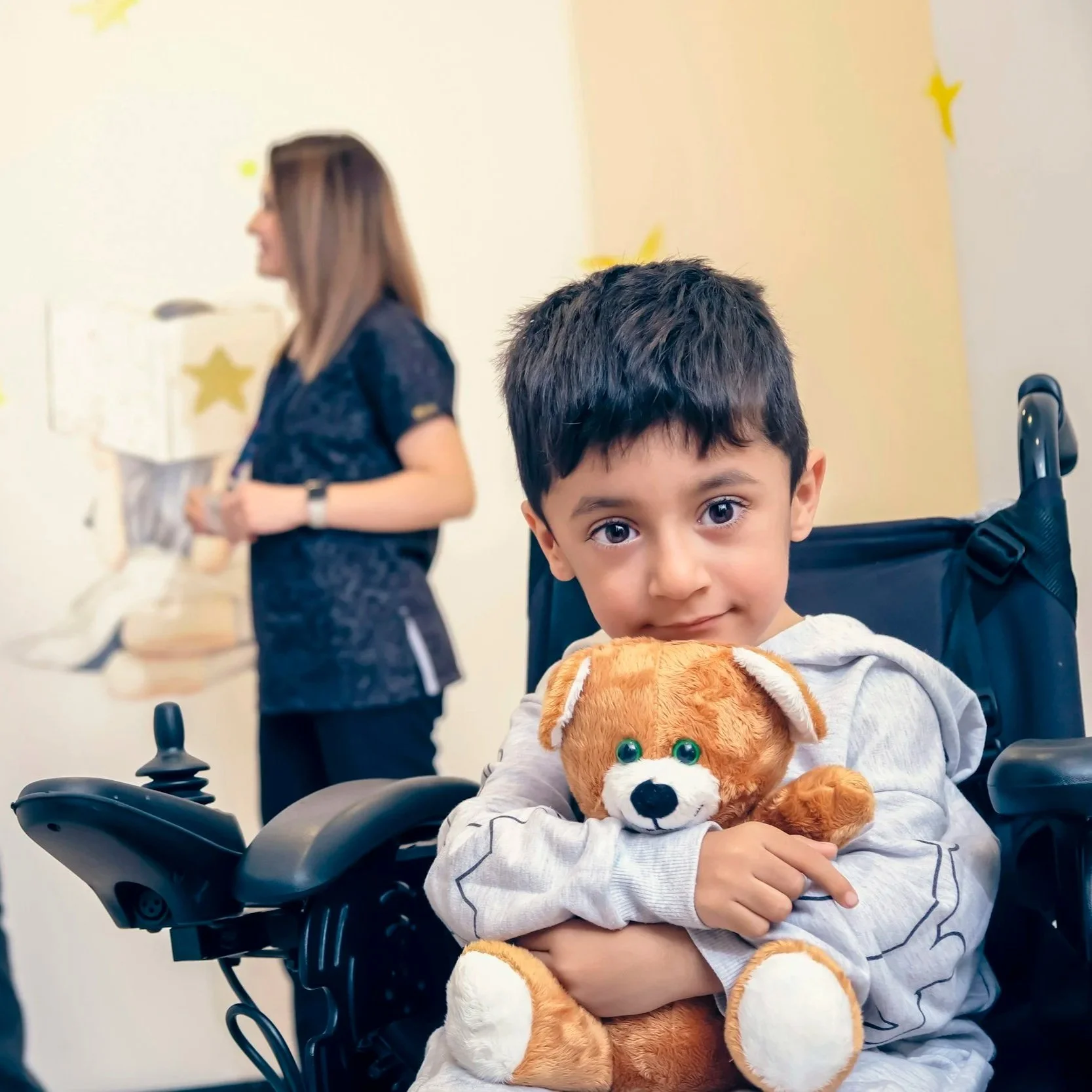 Bambino seduto in sedia a rotelle con un orsacchiotto, mentre una donna sullo sfondo tiene un vassoio con torte.