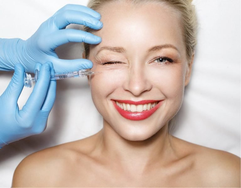 Donna mentre riceve un'iniezione estetica dal medico, che indossa guanti blu, con un sorriso e un trucco leggero.