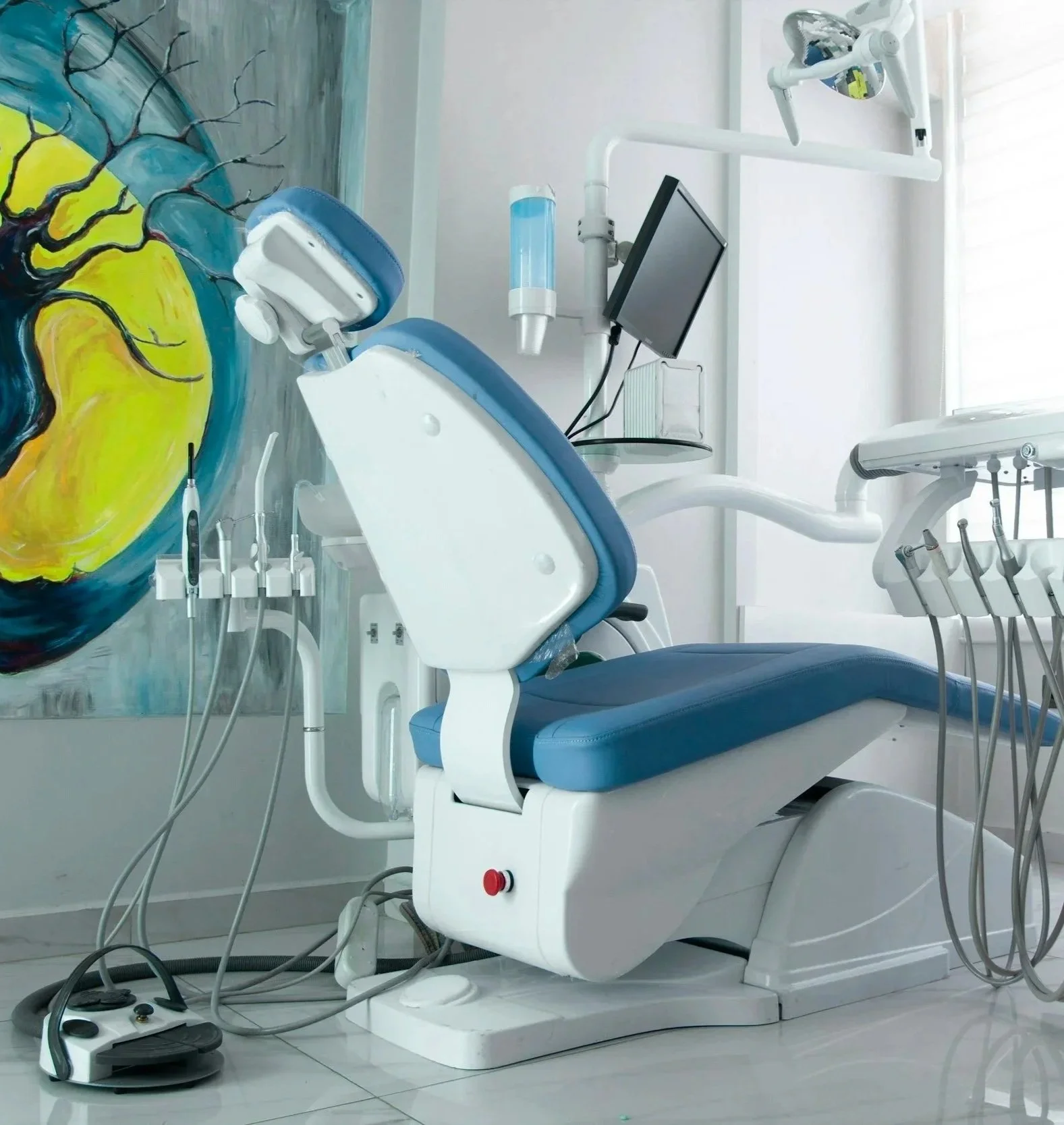 Sedia odontoiatrica con strumentazione dentale e monitor, parete decorata con murale astratto.