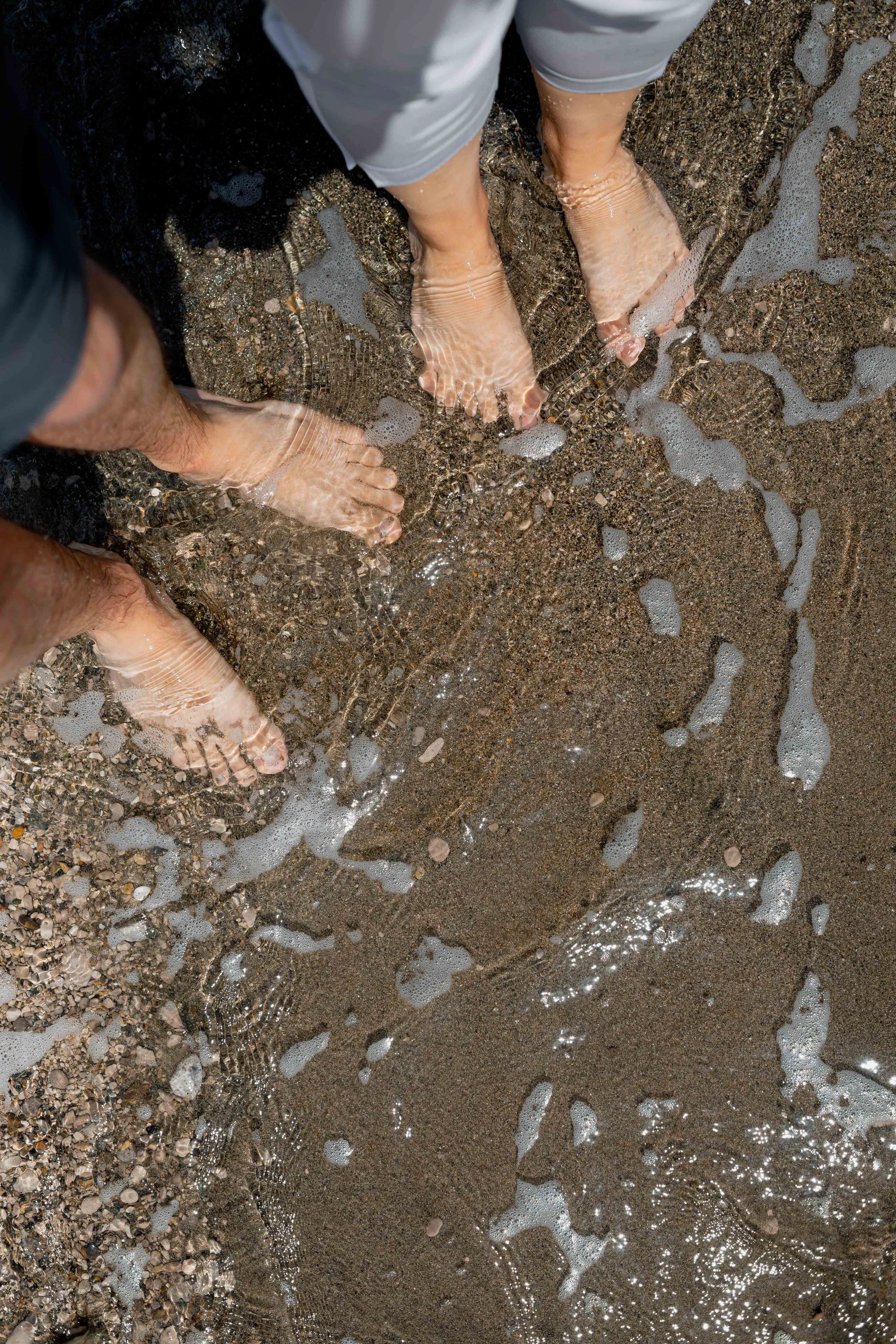 Two pairs of feet standing in shallow water on a sandy beach, with small waves and foam around them.
