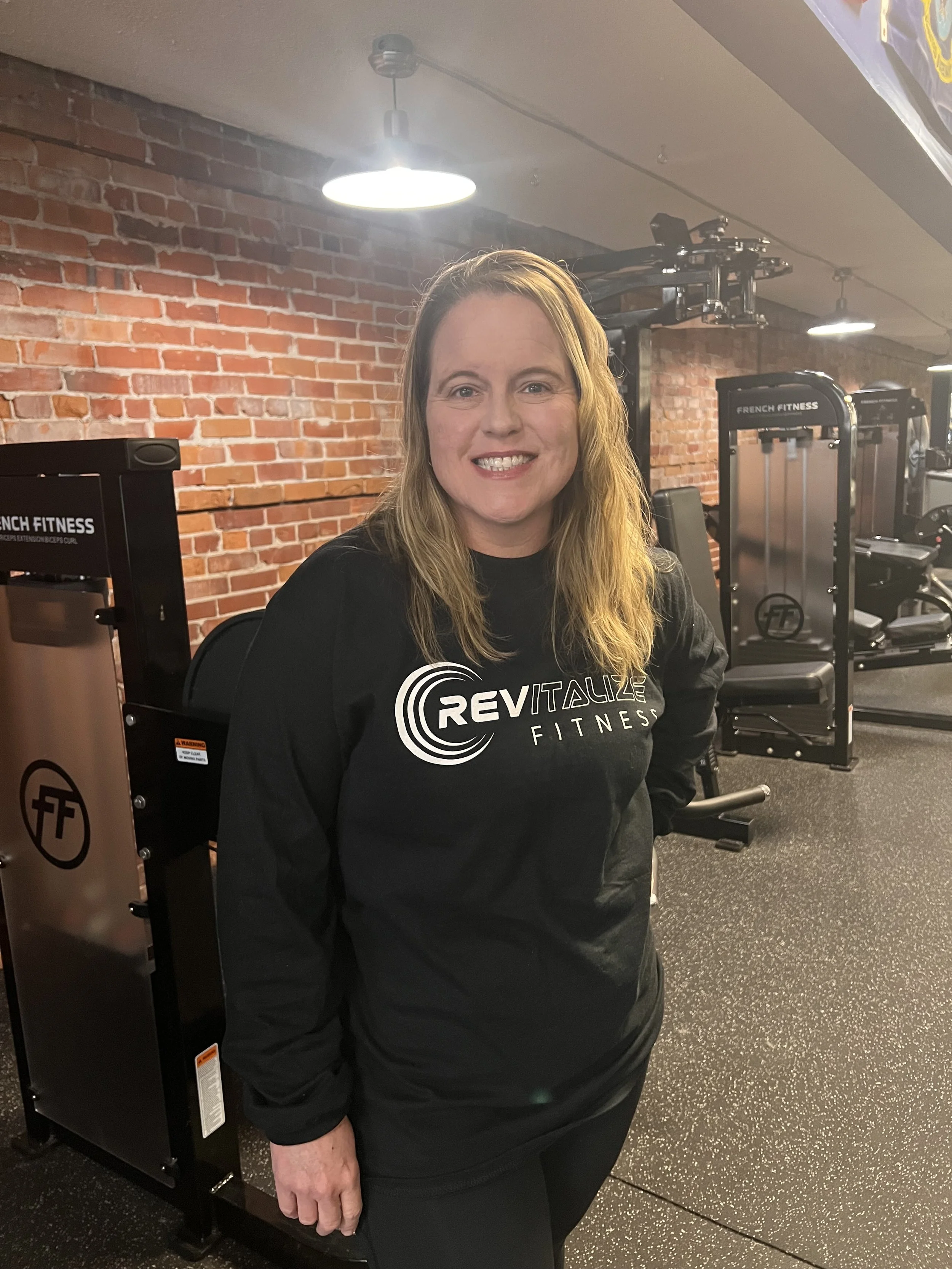 A woman standing in front of a blue shelf with nutrition supplements and a sign for Rick Anderson Fitness, wearing a black T-shirt with the same logo and text.