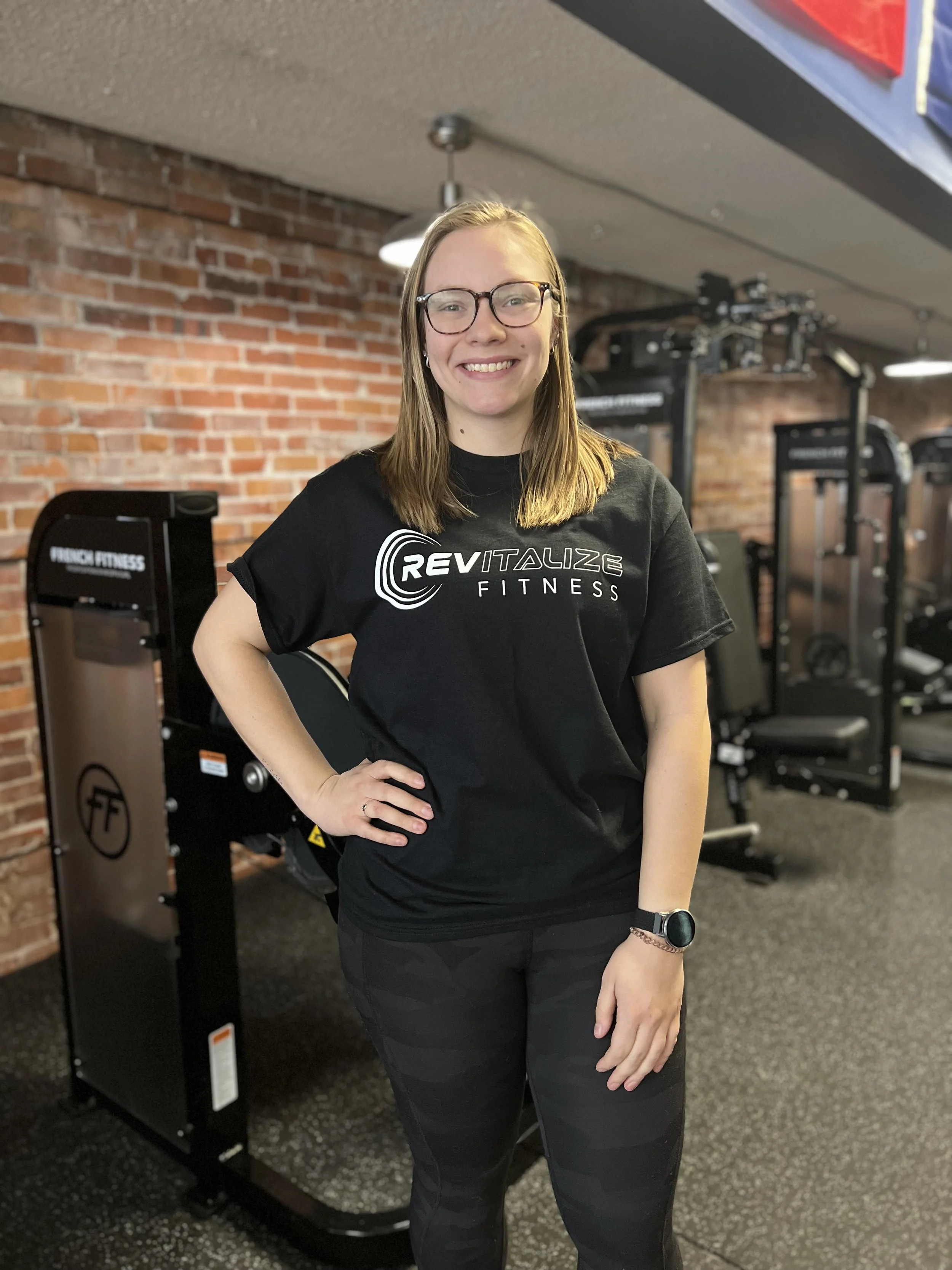 A young woman with glasses and long blonde hair smiling at the camera in a gym, wearing black workout clothing, with workout equipment and a brick wall in the background.