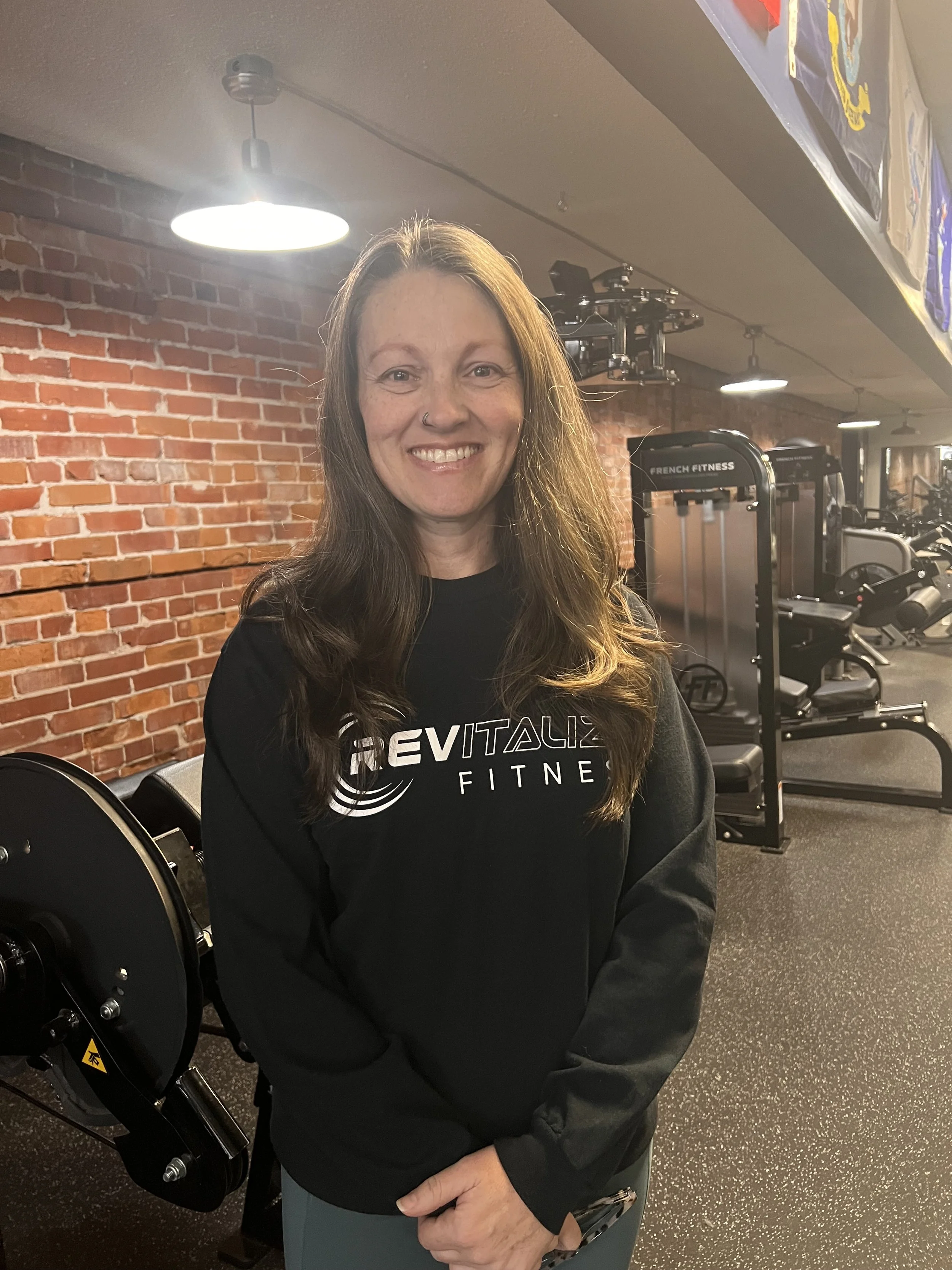 A woman smiling in front of a Rick Anderson Fitness sign, wearing a black T-shirt with the company logo and text for personal training, nutrition coaching, and fitness.