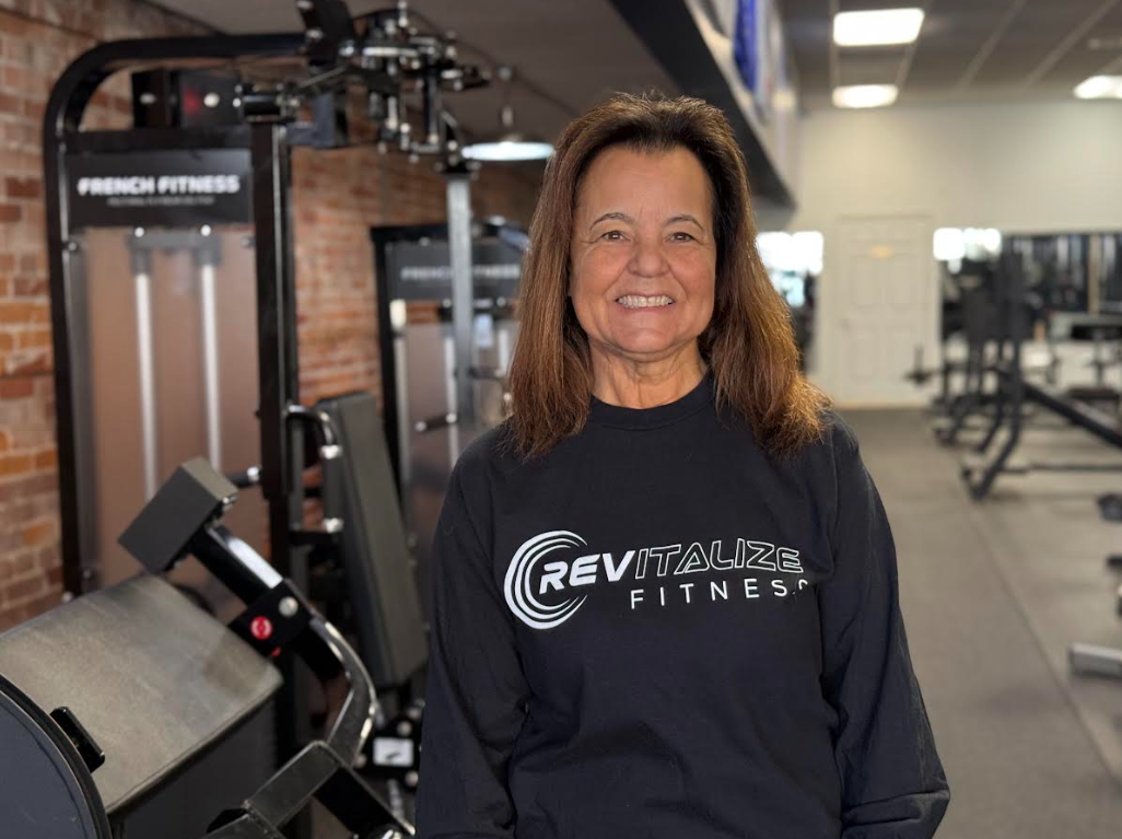 A smiling middle-aged woman with shoulder-length brown hair, wearing a black T-shirt with the logo 'RA' and red pants, standing inside a gym with American flags and fitness equipment in the background.