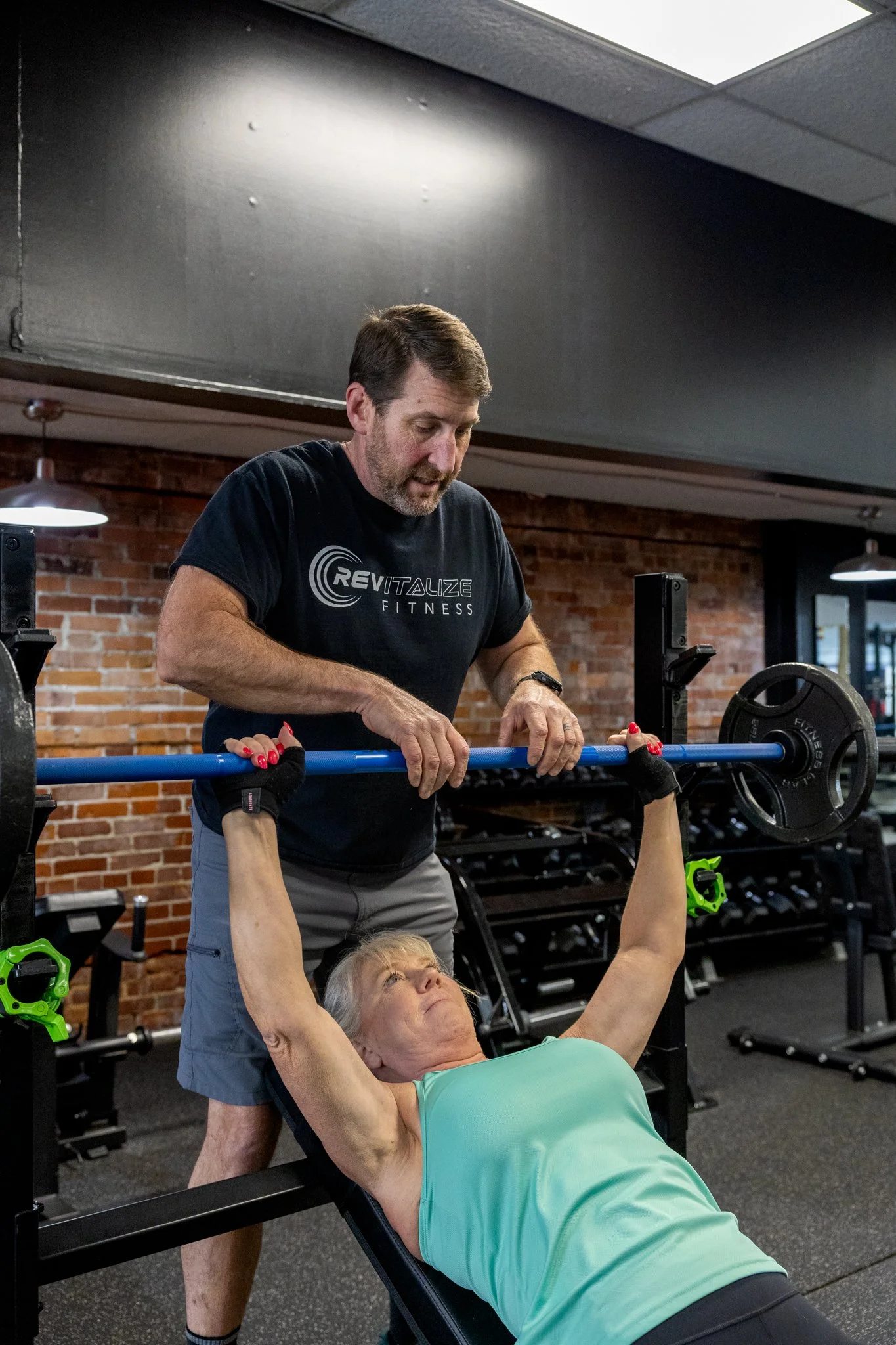 A woman lifting a dumbbell with guidance from a trainer inside a gym.