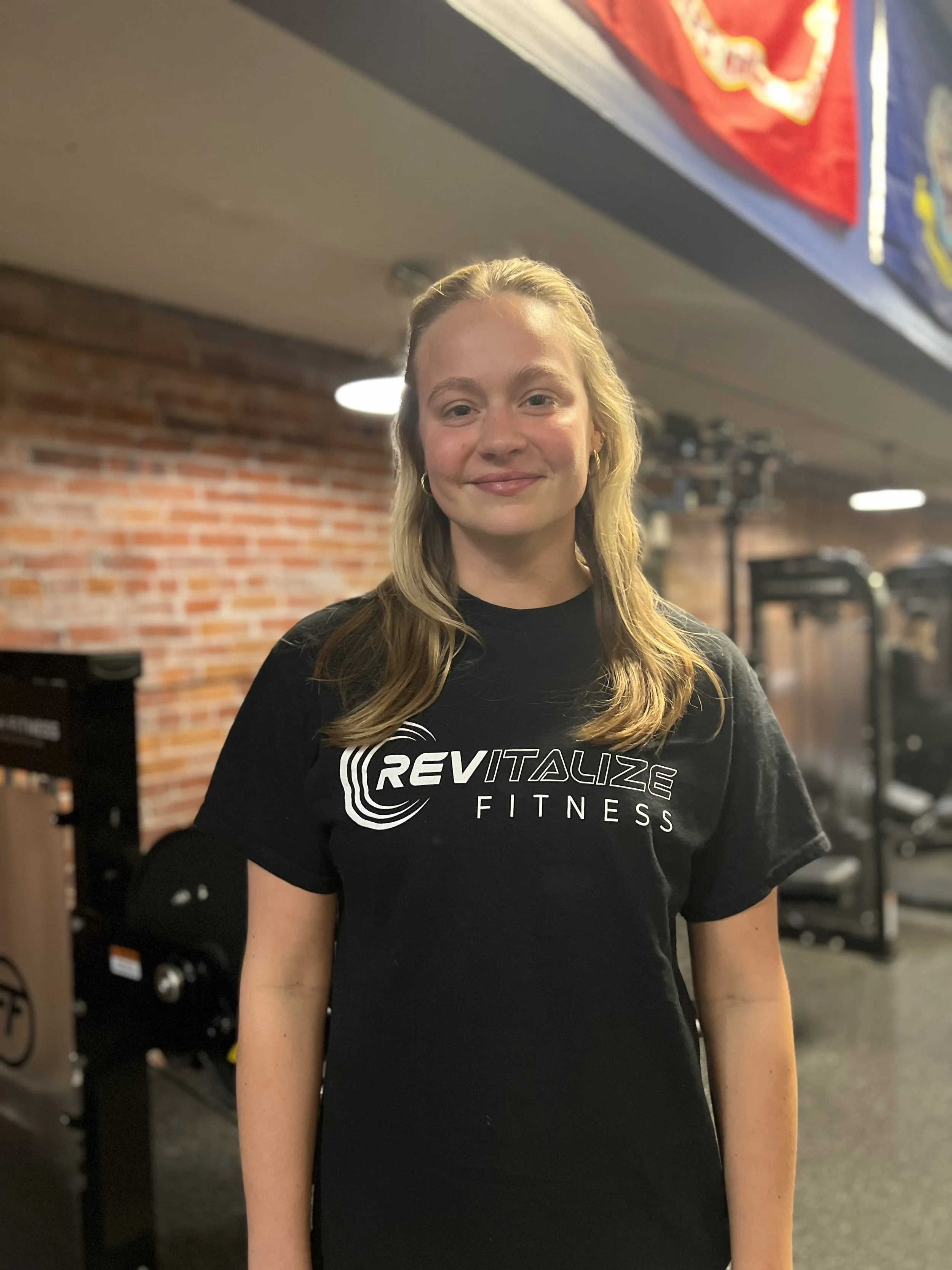 A young woman with blonde hair smiling, standing in front of a fitness nutrition shop with a sign that reads 'Rick Anderson Fitness Personal Nutrition'. She is wearing a black T-shirt with the same logo and text.