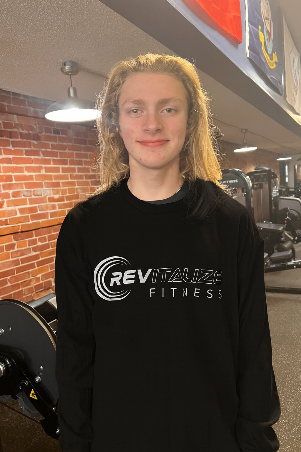 A young man standing in front of a teal-colored counter with a sign for Rick Anderson Fitness. He is wearing a black T-shirt with the company's logo and text, and smiling slightly. The background features fitness-related items and branding, including bottles and jars on shelves.