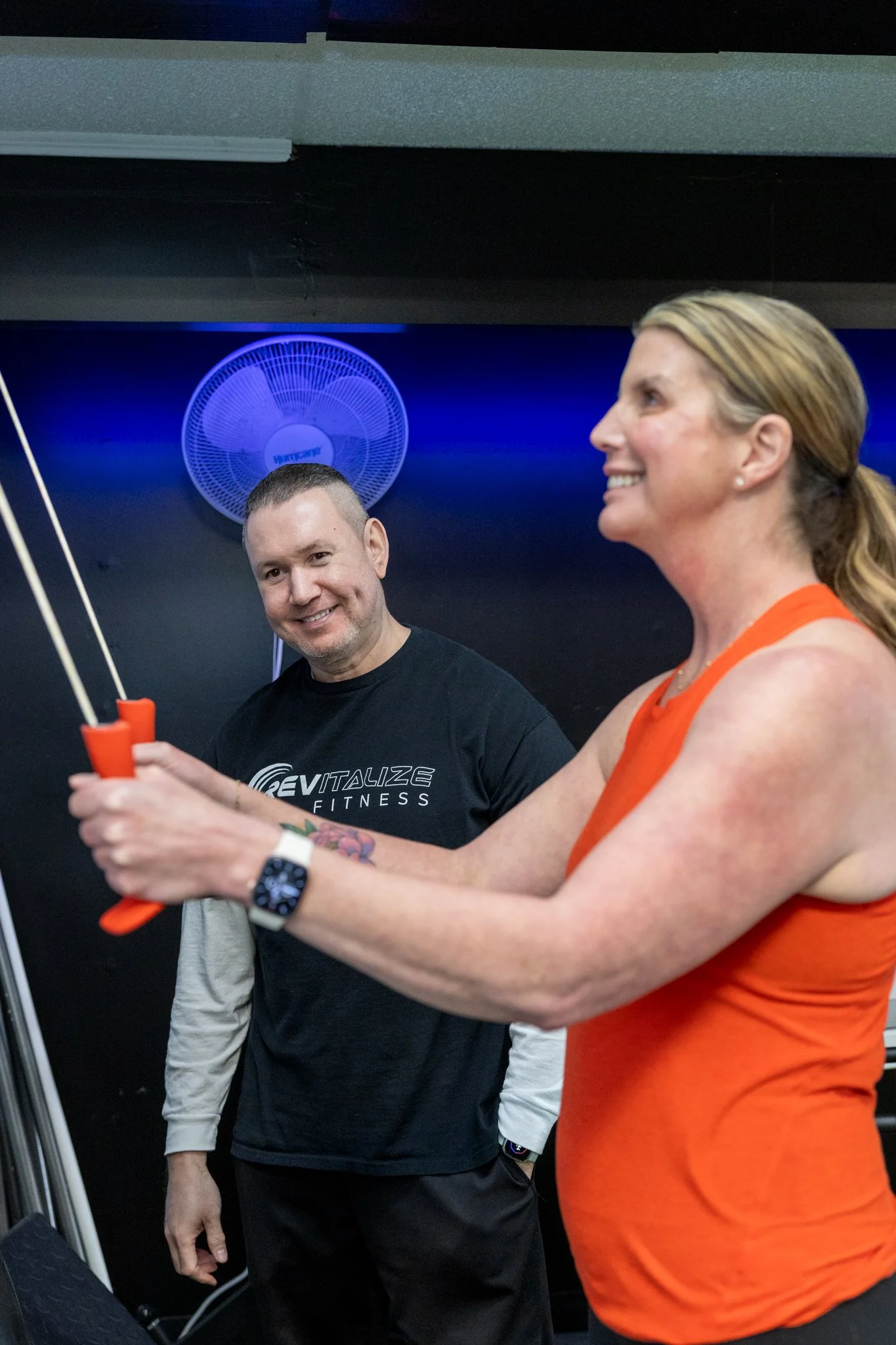 A woman is seated on gym equipment while a trainer provides instructions in a gym setting.